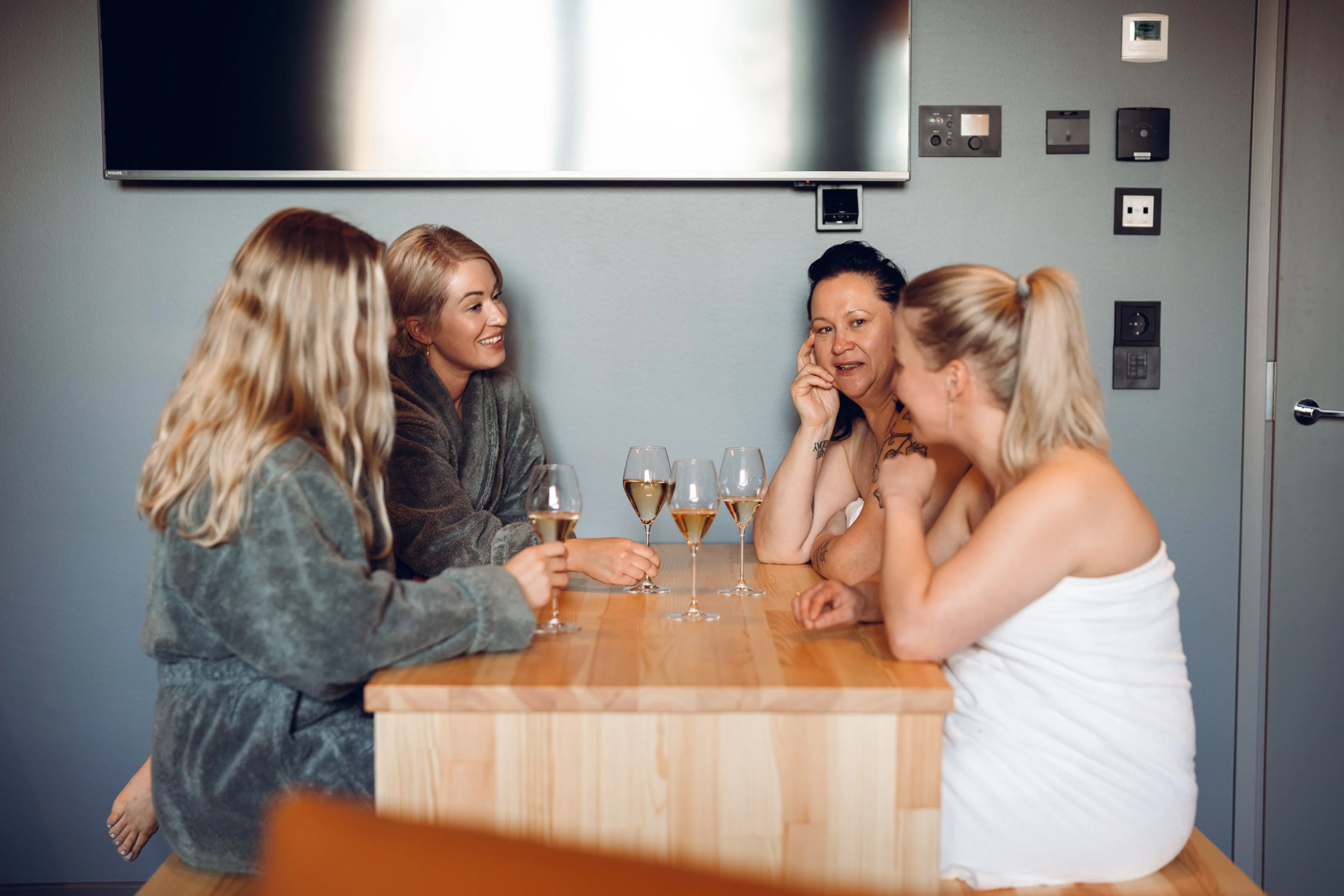 Neljä henkilöä saunaviittoihin ja pyyhkeisiin kietoutuneina nauttimassa viinilaseista puisen pöydän ääressä.