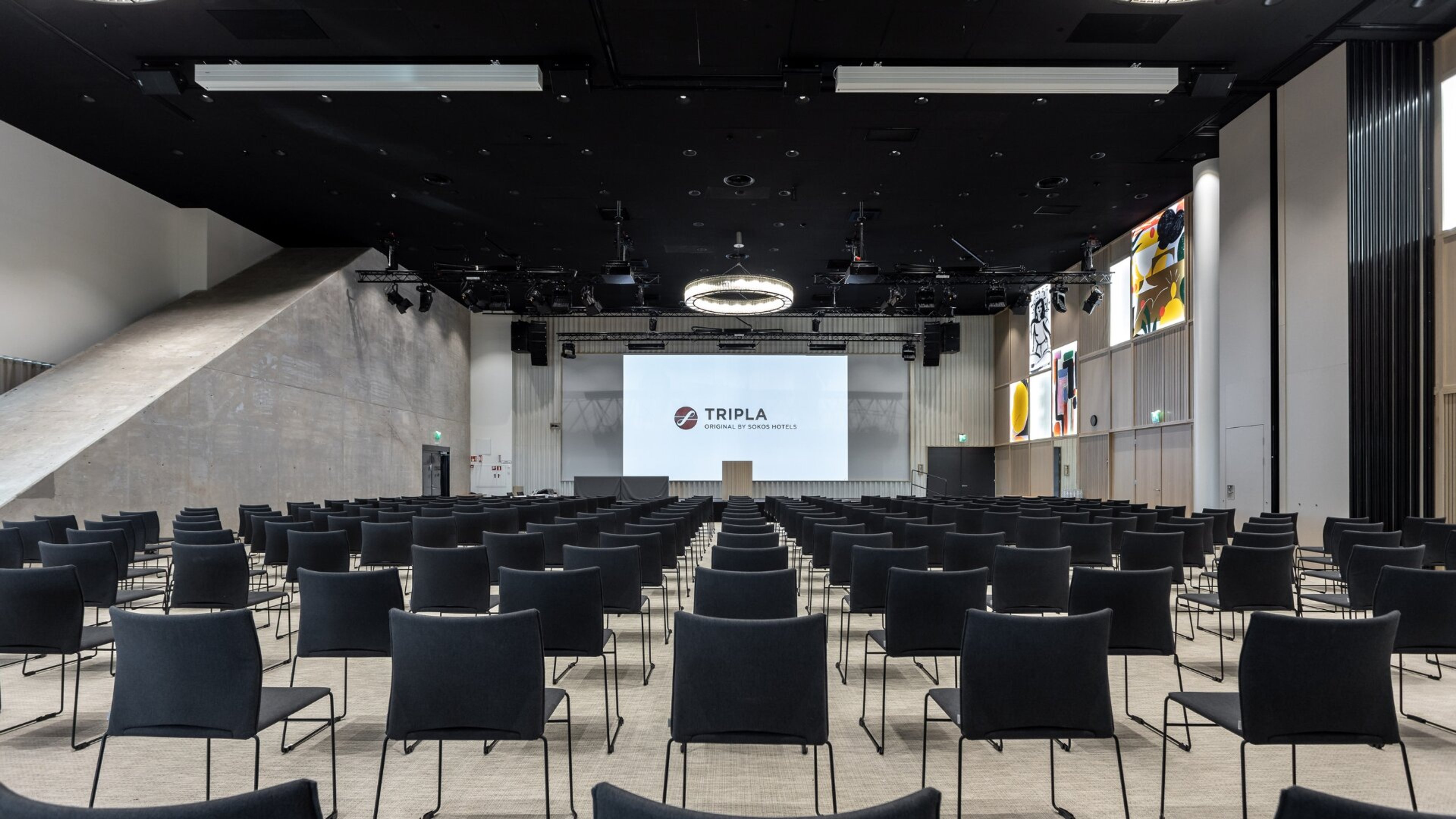 Moderni auditorio, jossa on rivit sinisiä tuoleja, lavalla valkoinen projektorikangas, korkeat betoniset seinät ja musta kattopinta.