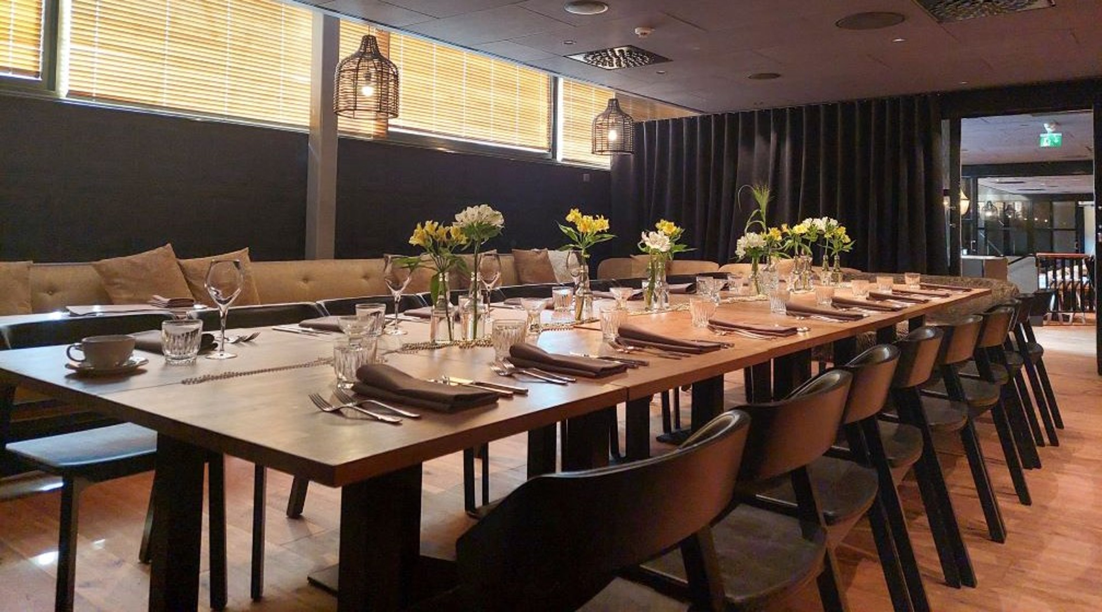 Elegantti ravintolan yksityistila, jossa pitkä puinen pöytä katettuna, kukka-asetelmia, mukavat tuolit ja sohva taustalla. Kovaa puulattiaa.