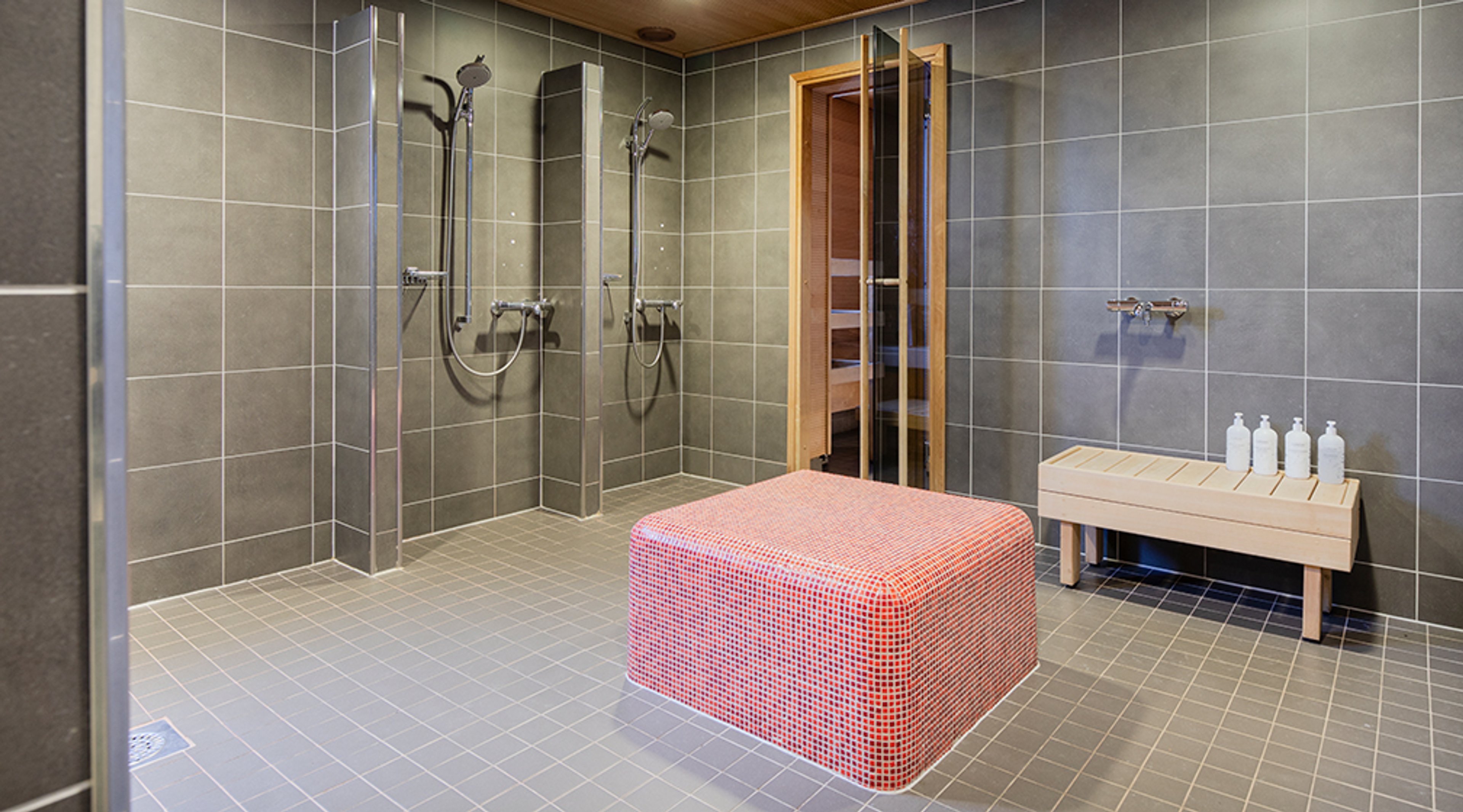 Moderni saunaosasto harmain laatoitetuin seinin, kaksi suihkua, punainen tasonpäällinen istuinpenkki keskellä ja puinen ovi saunaan.