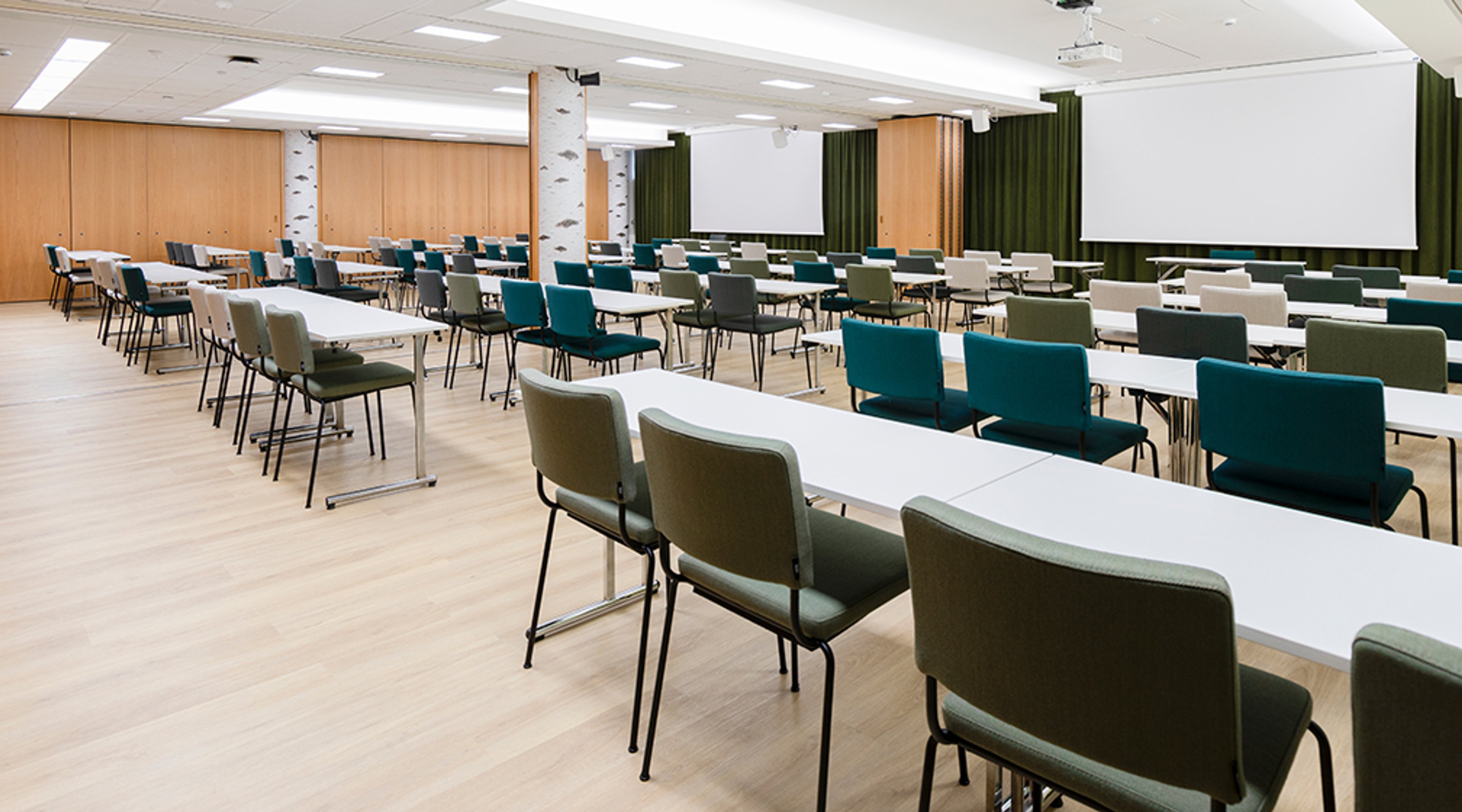 Moderni kokoustila valkoisilla pöydillä ja sinisillä, vihreillä sekä harmailla tuoleilla. Takaseinällä projektorikangas ja vihreät verhot.