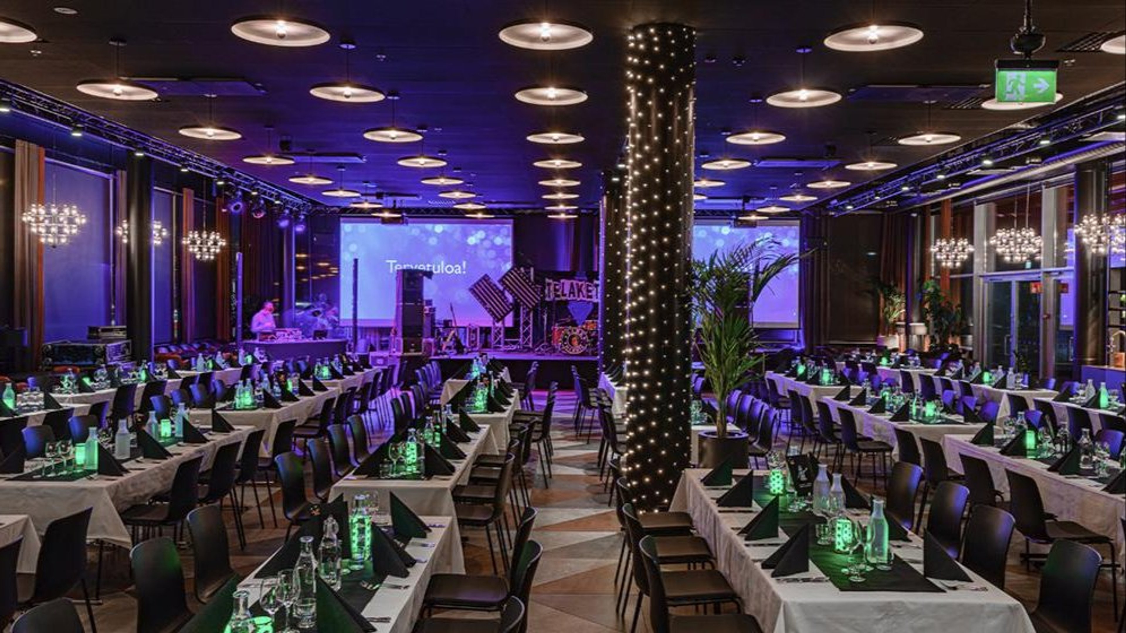 Tilava auditorio, jossa pöydät valmiina tilaisuutta varten. Violetti valaistus, suuri valkokangas edessä ja tasainen kova lattia pitkillä pöytäriveillä.