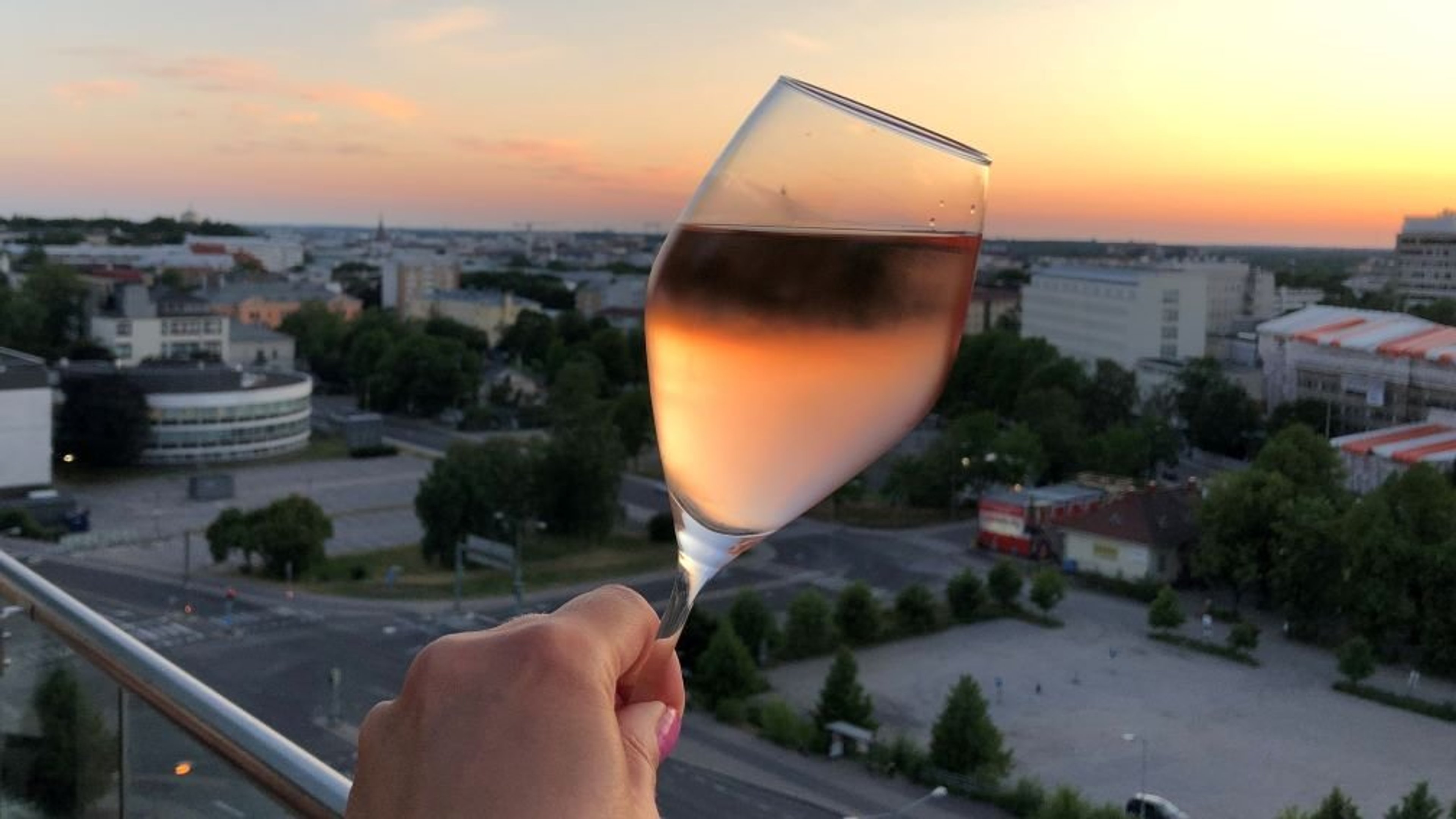 Samppanjalasi kädessä, taustalla kaupunkimaisema auringonlaskun värjäämänä oranssiksi ja vaaleanpunaiseksi.