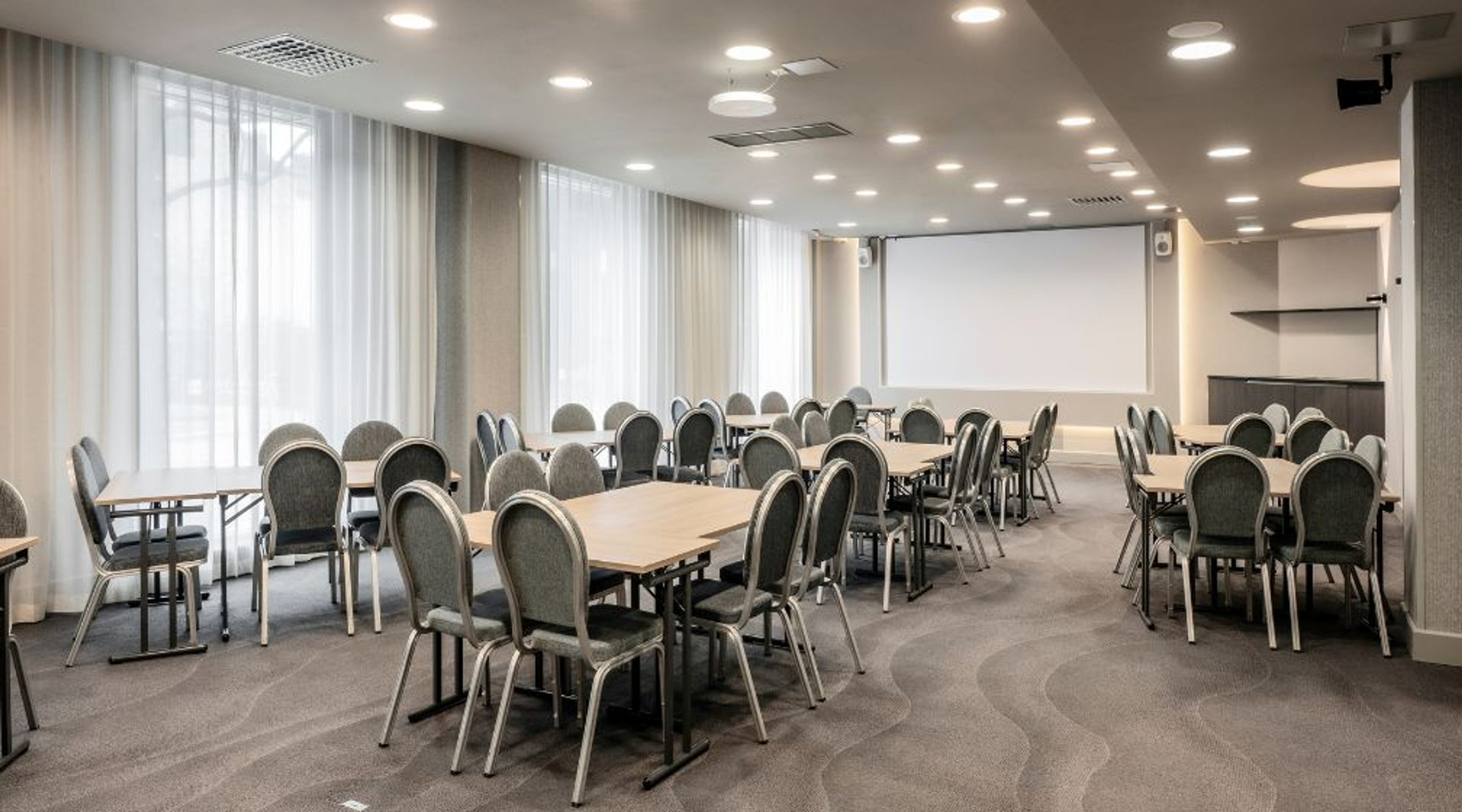 Valoisa kokoushuone useilla pöydillä ja tuoleilla. Isot ikkunat, kokolattiamatto ja valkoinen projektiokangas edessä.