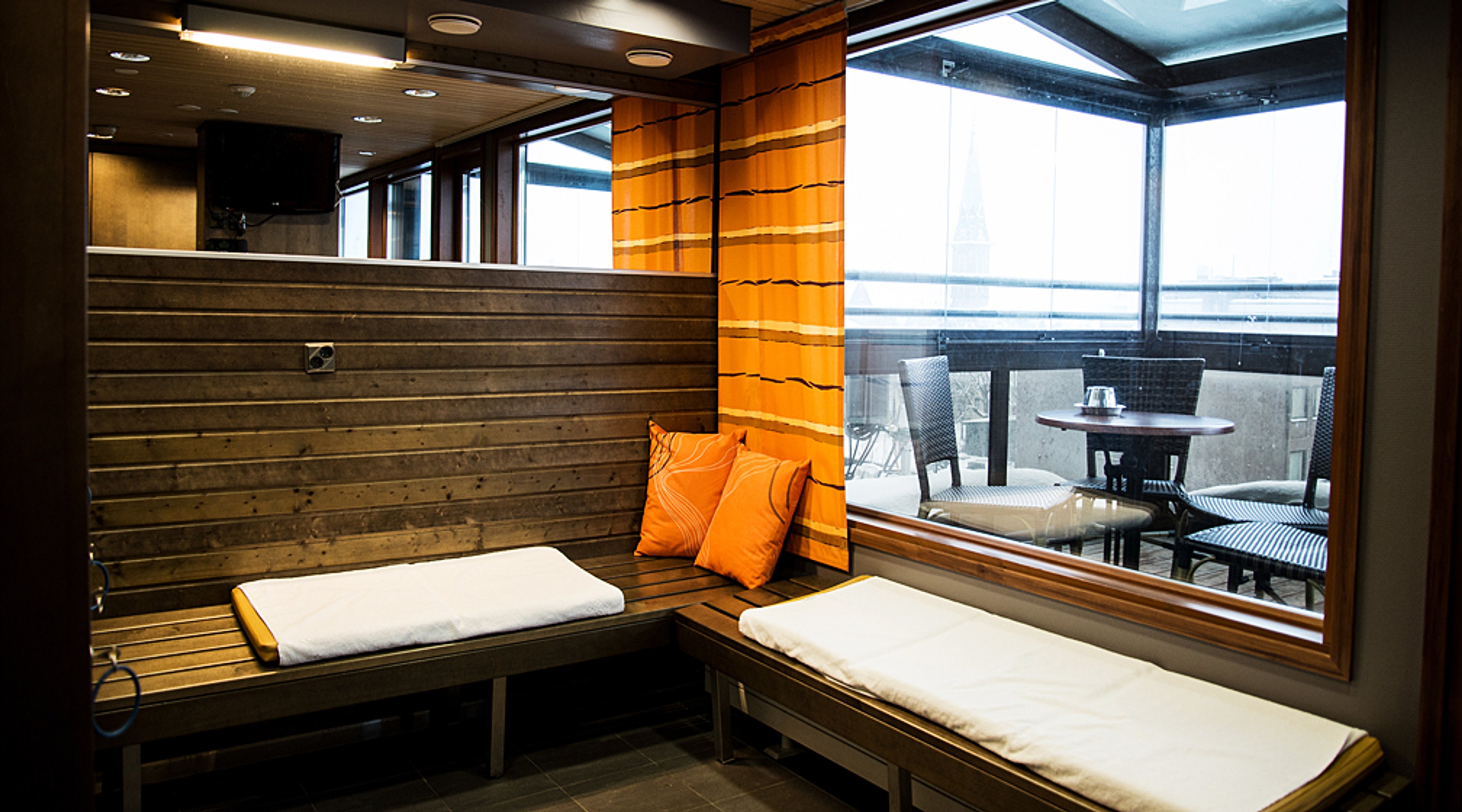Sauna, jossa puinen seinäpanelointi, oranssit tyynyt ja valkoiset pyyhkeet. Ikkunan takana näkyy ulkoterassi pöydillä.