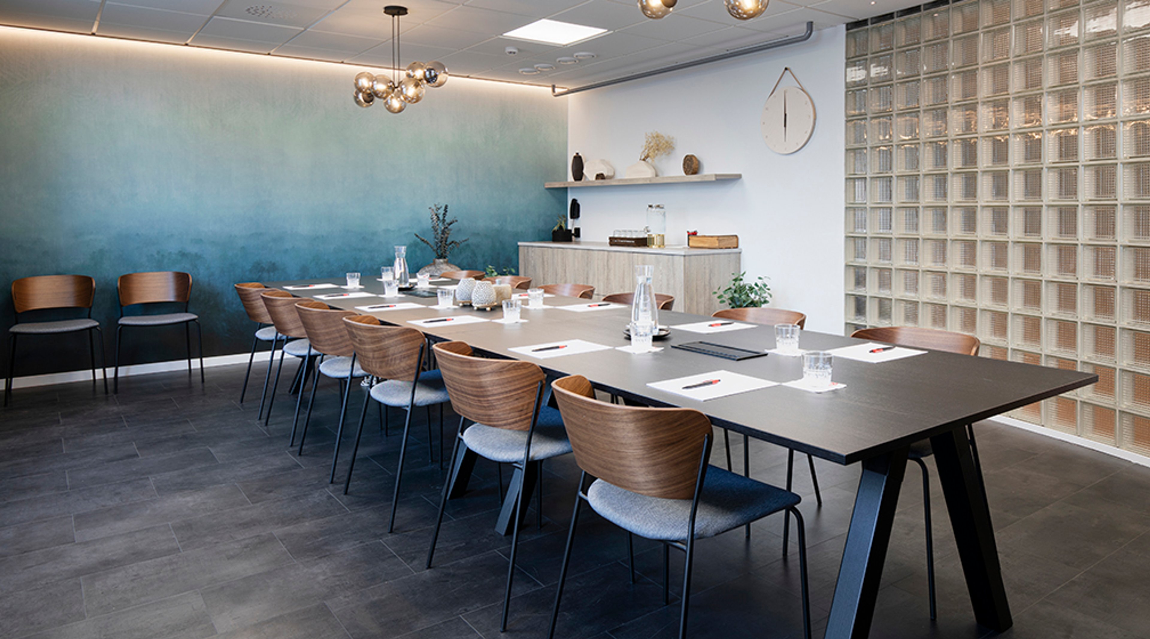 Moderni kokoushuone siniturkoosilla seinällä. Pitkän pöydän ympärillä puuselkänojallisia tuoleja. Kova laattalattia ja lasinen seinäelementti.