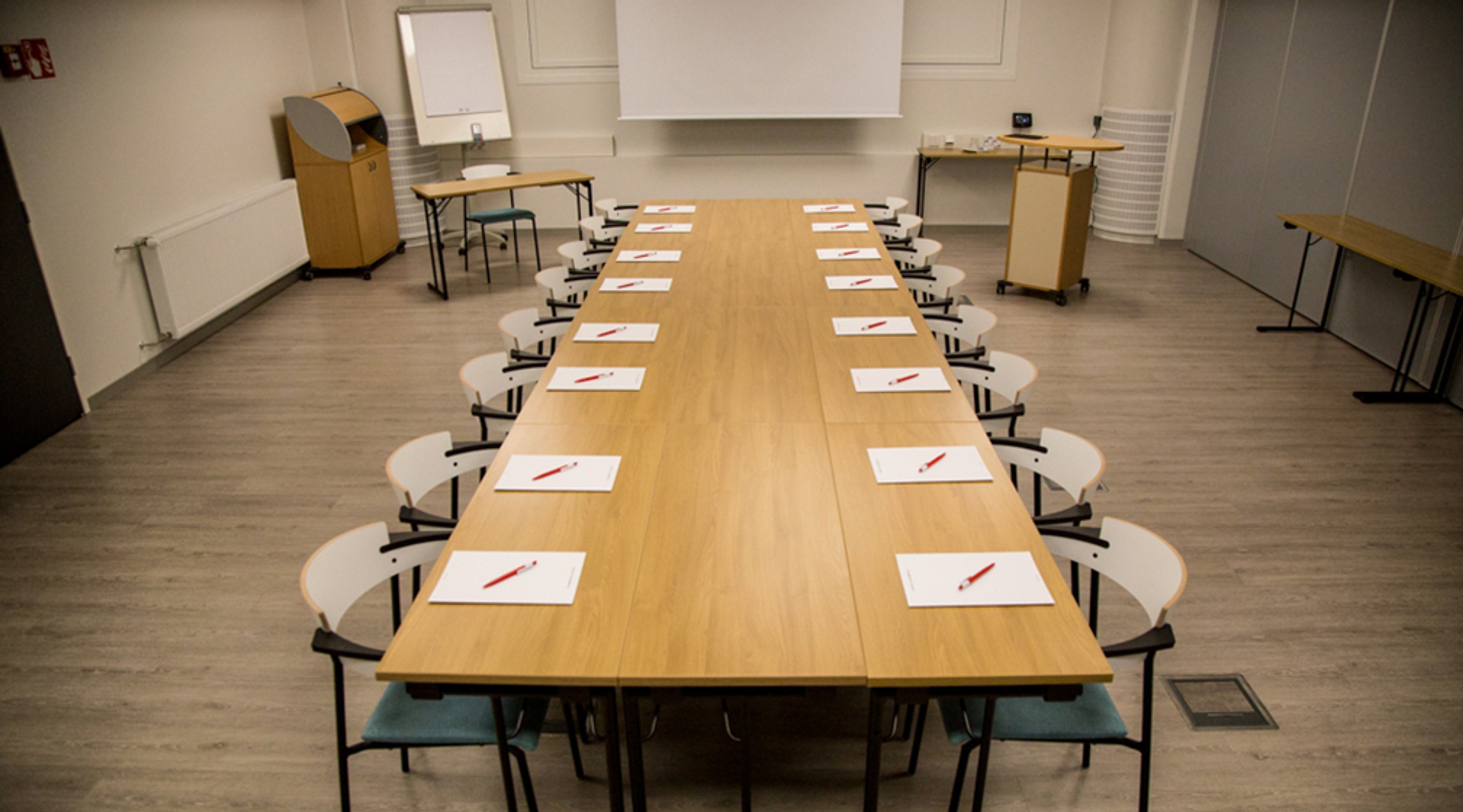 Kokoushuone, jossa pitkä puinen pöytä, valkoisia tuoleja ja muistiinpanovälineitä. Tilassa on puulattia ja valkokangas.