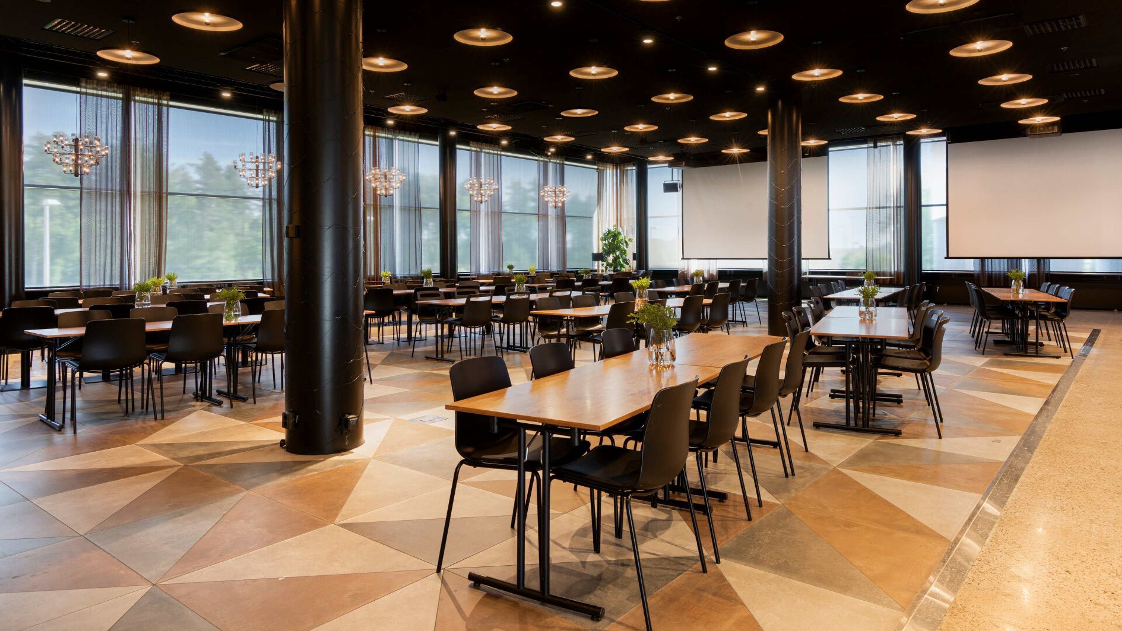 Kaasaegne konverentsiruum laudade ja toolidega. Mustrilise põranda, ümmarguste laevalgustite ja suured aknad metsavaatega. Ruumis on projektoriekraanid.