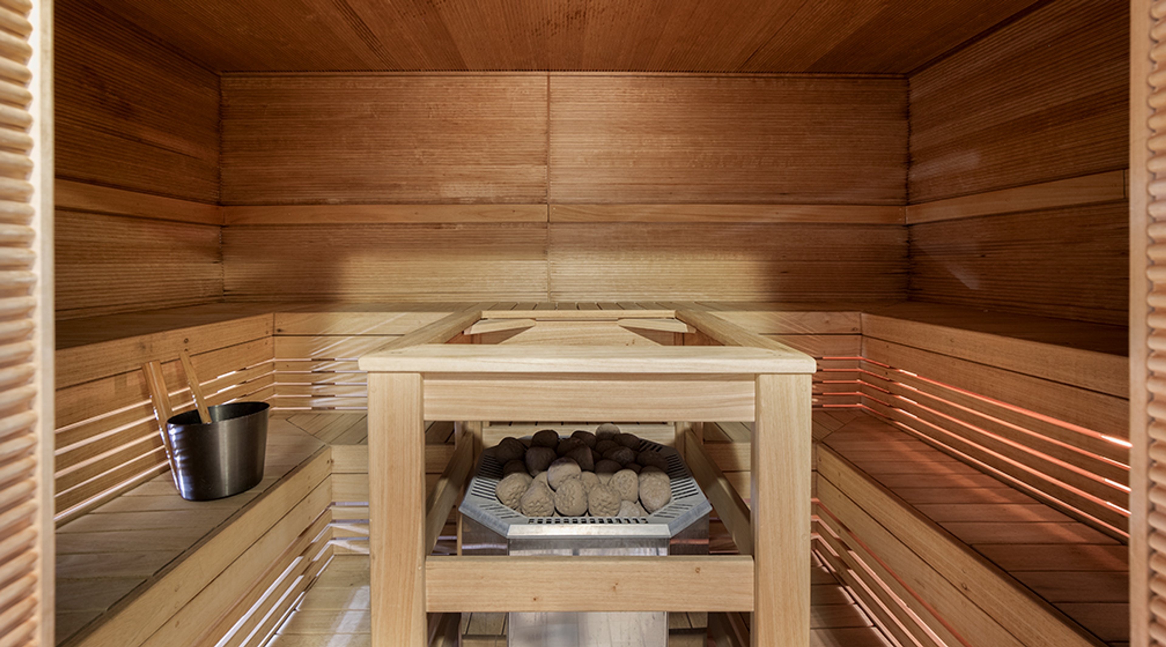 Tyylikkään saunan sisätila puupaneelilla. Keskellä kiuas, jossa kiviä. Lauteilla vesiämpäri ja kauha.