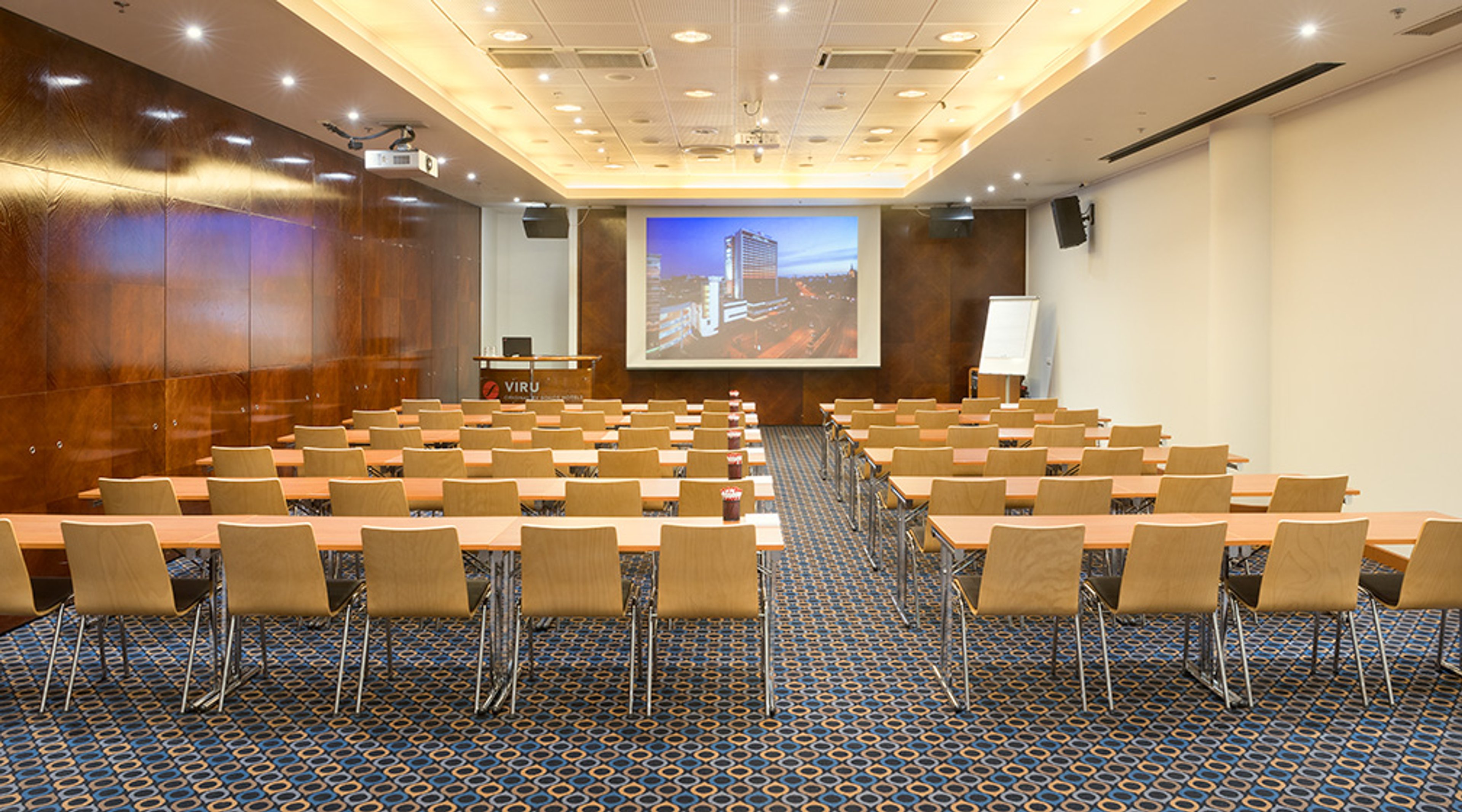 Moderni auditoriotila ruskein puupaneelein, vaaleilla tuoleilla ja sinisellä kuvioidulla matolla. Edessä suuri valkokangas ja luentopöytä.