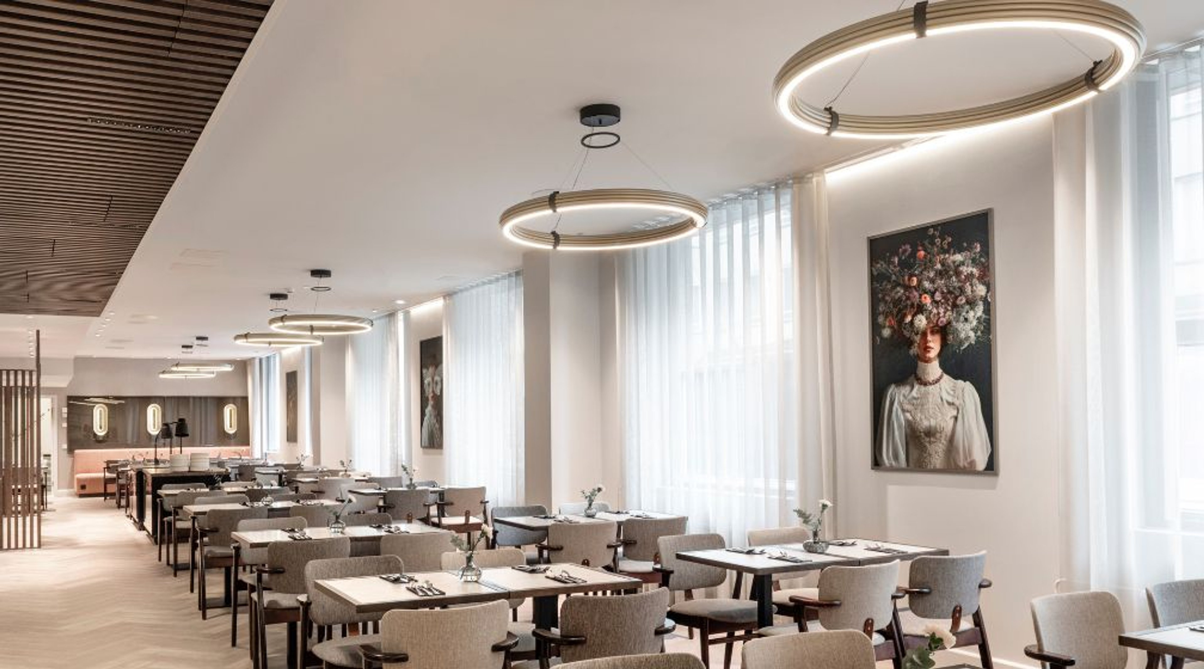 Valoisa ravintola, jossa pyöreät valaisimet katossa, harmaat tuolit ja valkoisia pöytiä. Seinällä kukka-aiheisia taideteoksia.