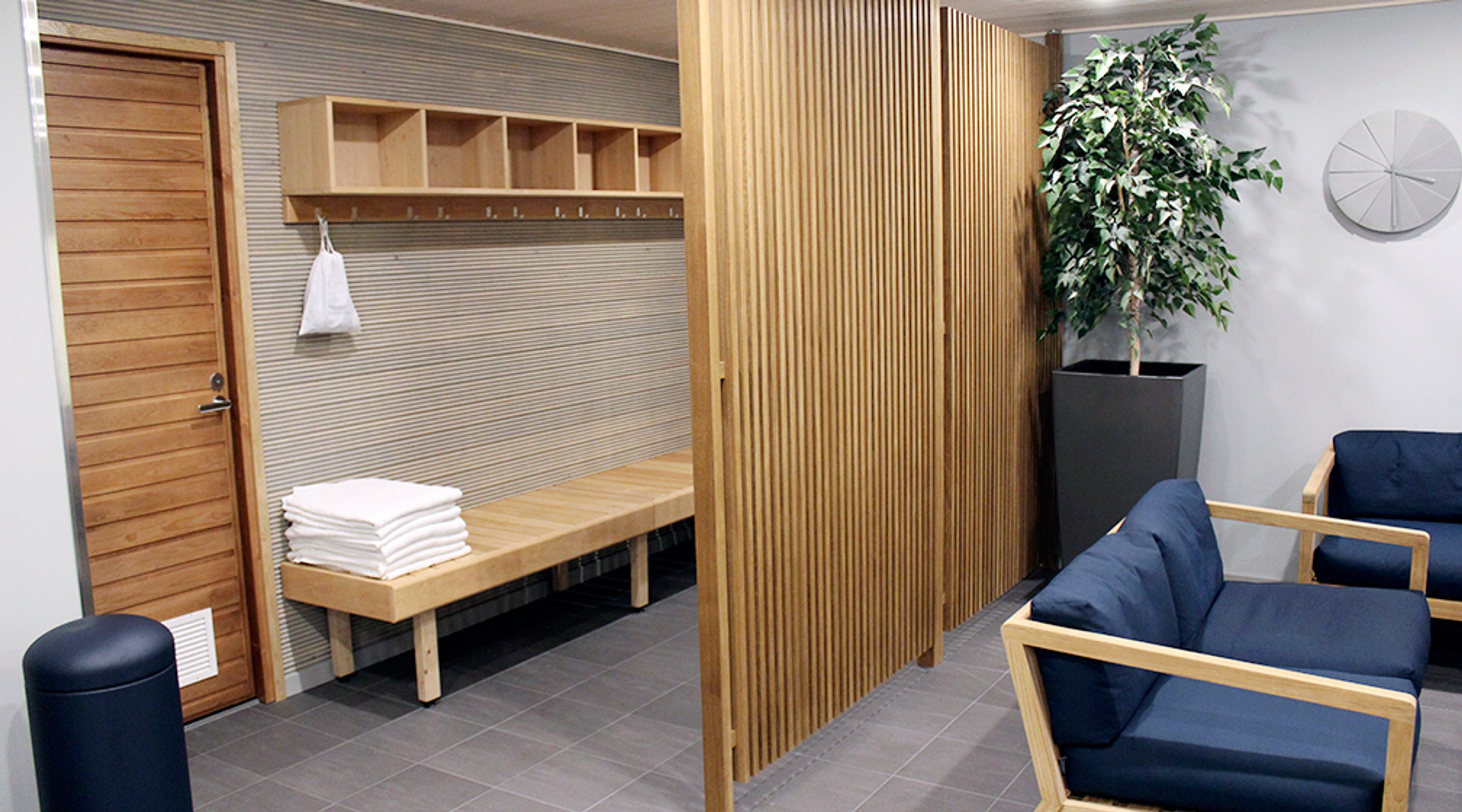 Saunan pukuhuone puuseinällä jaettuna. Penkki, pyyhkeitä, naulakko, sininen sohva ja viherkasvi. Harmaa laattalattia.