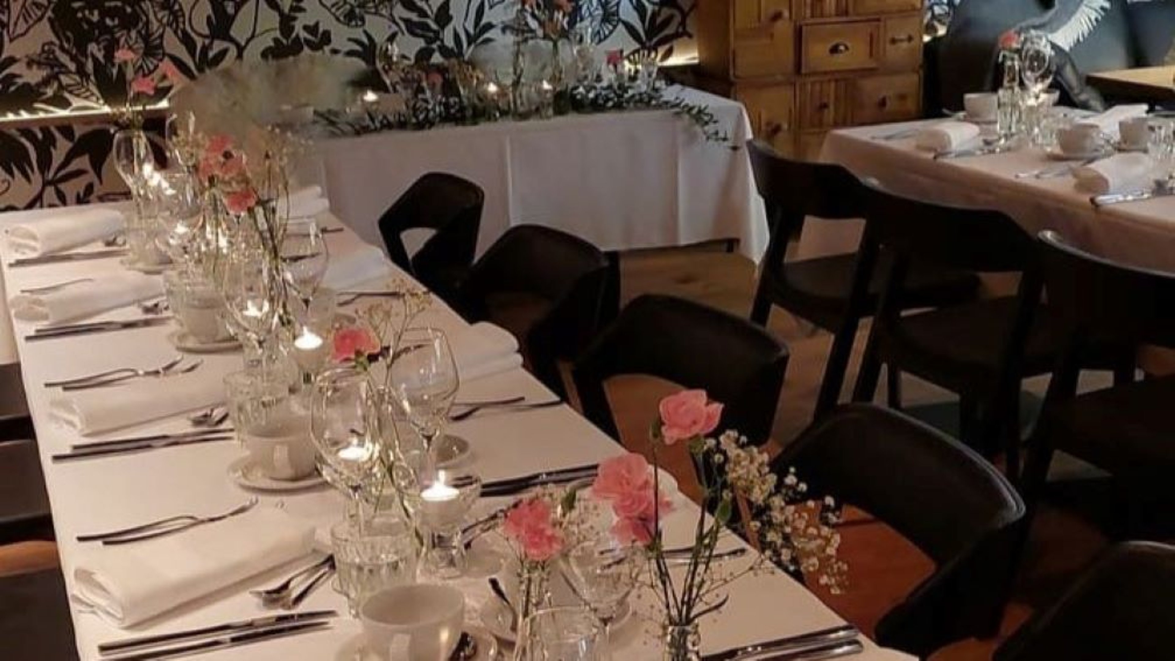 Ravintolan sisätila jossa pitkät pöydät valkoisilla liinoilla, mustilla tuoleilla, koristeena vaaleanpunaisia kukkia ja kynttilöitä.
