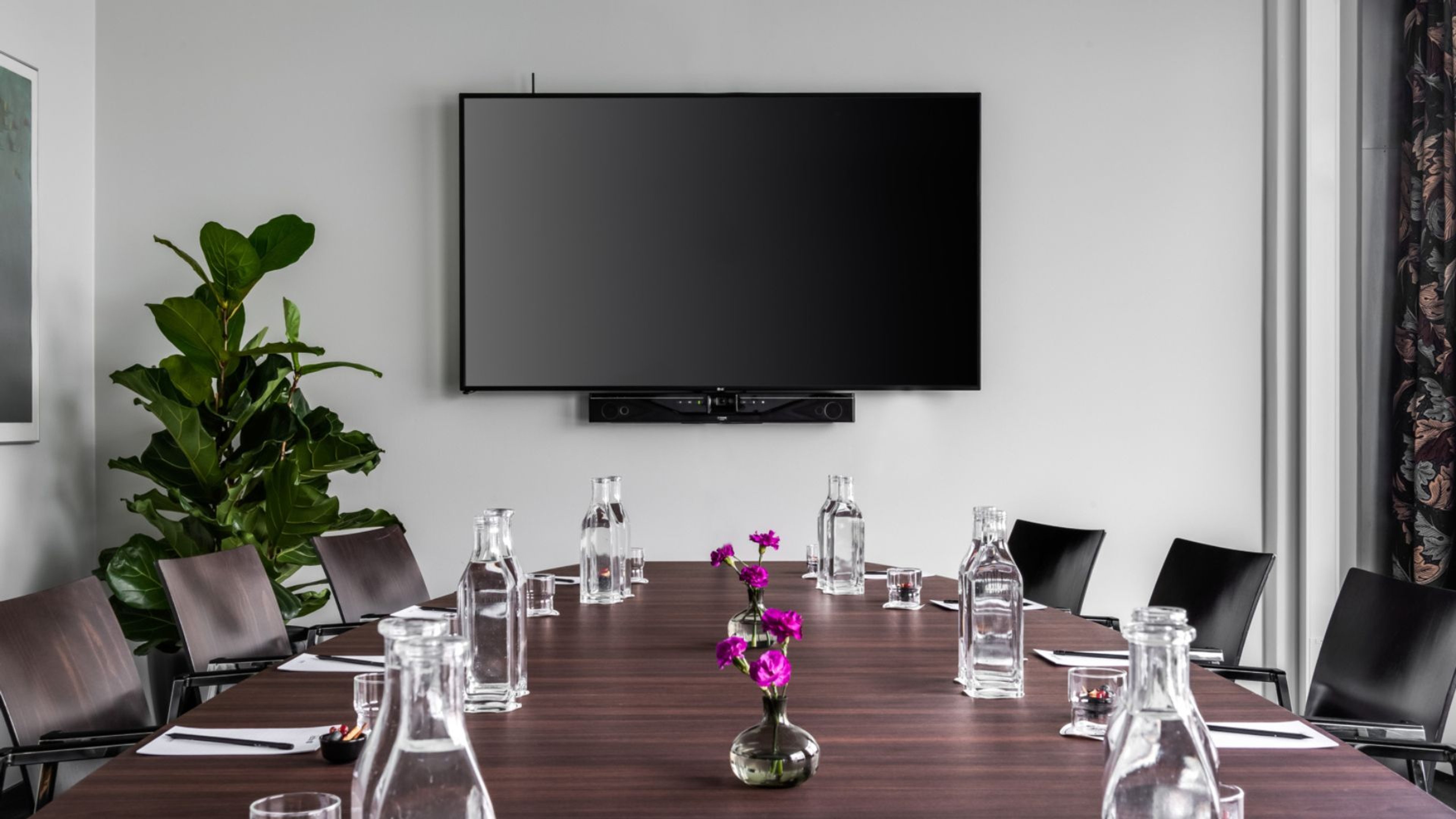 Kokoushuone, jossa tumma puinen pöytä, lasipulloja, tuoleja ja seinään kiinnitetty televisio. Vieressä viherkasvi ja pöydällä violetteja kukkia.
