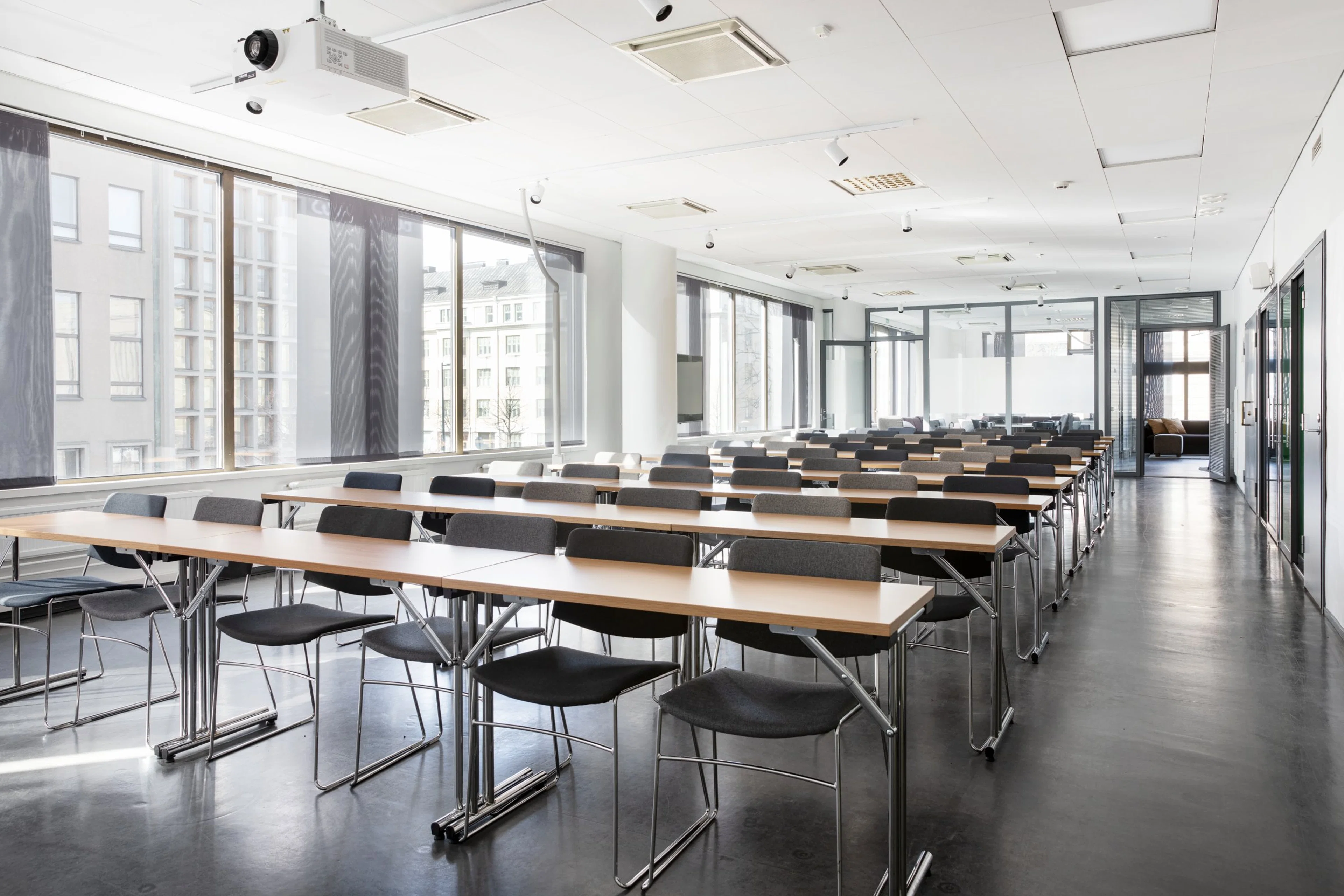 Valoisa luokkahuone pitkillä pöydillä ja tuoleilla. Isot ikkunat, joista näkyy kaupunkimaisema. Tasainen lattiapinta ja esteetön tila.