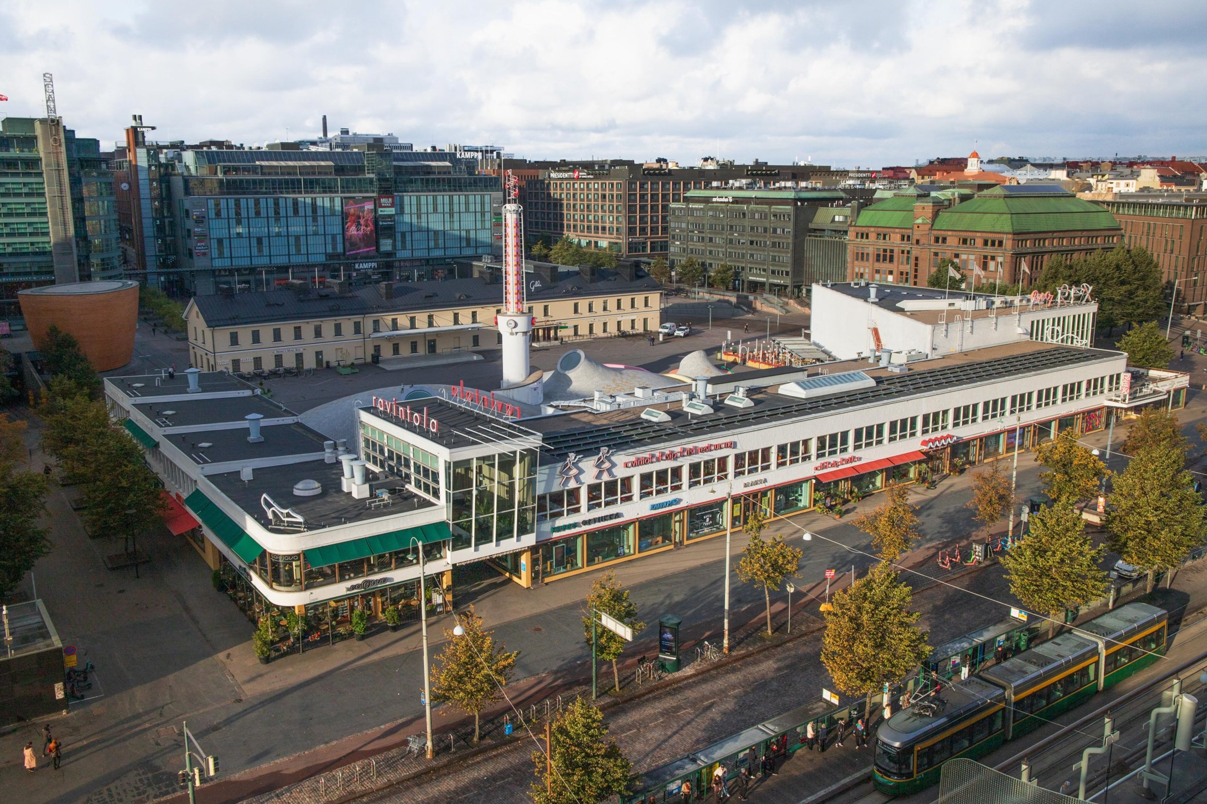 Flygbild över Glaspalatset och Kampens köpcentrum i centrala Helsingfors. En spårvagn kör i förgrunden med stadsvyn i bakgrunden.