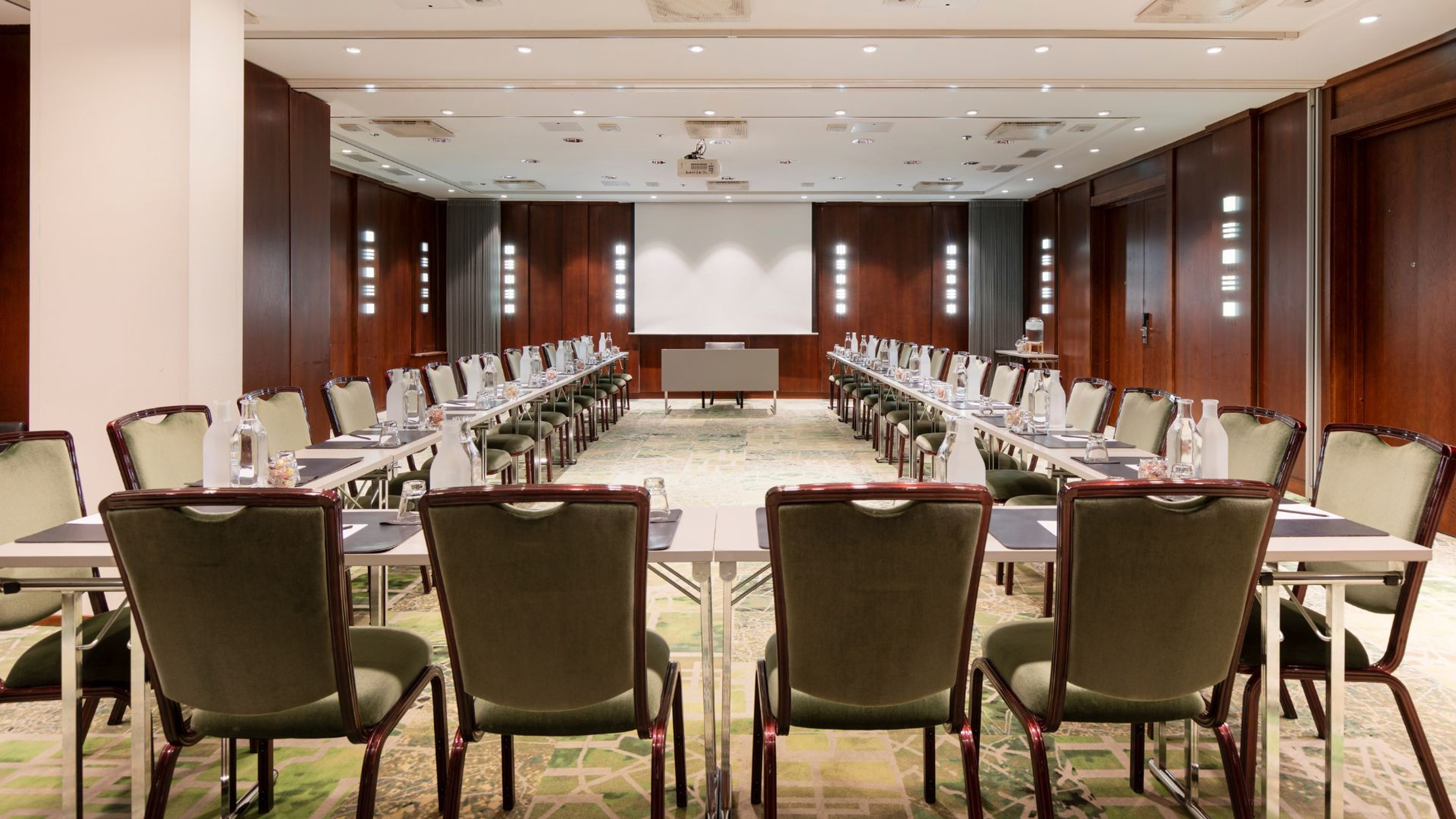 Kokoushuone U-muotoisella pöytäjärjestyksellä. Tummapaneloitu tila, vaaleat tuolit, valkokankaan edessä puhujanpönttö ja tasainen lattiapinta.