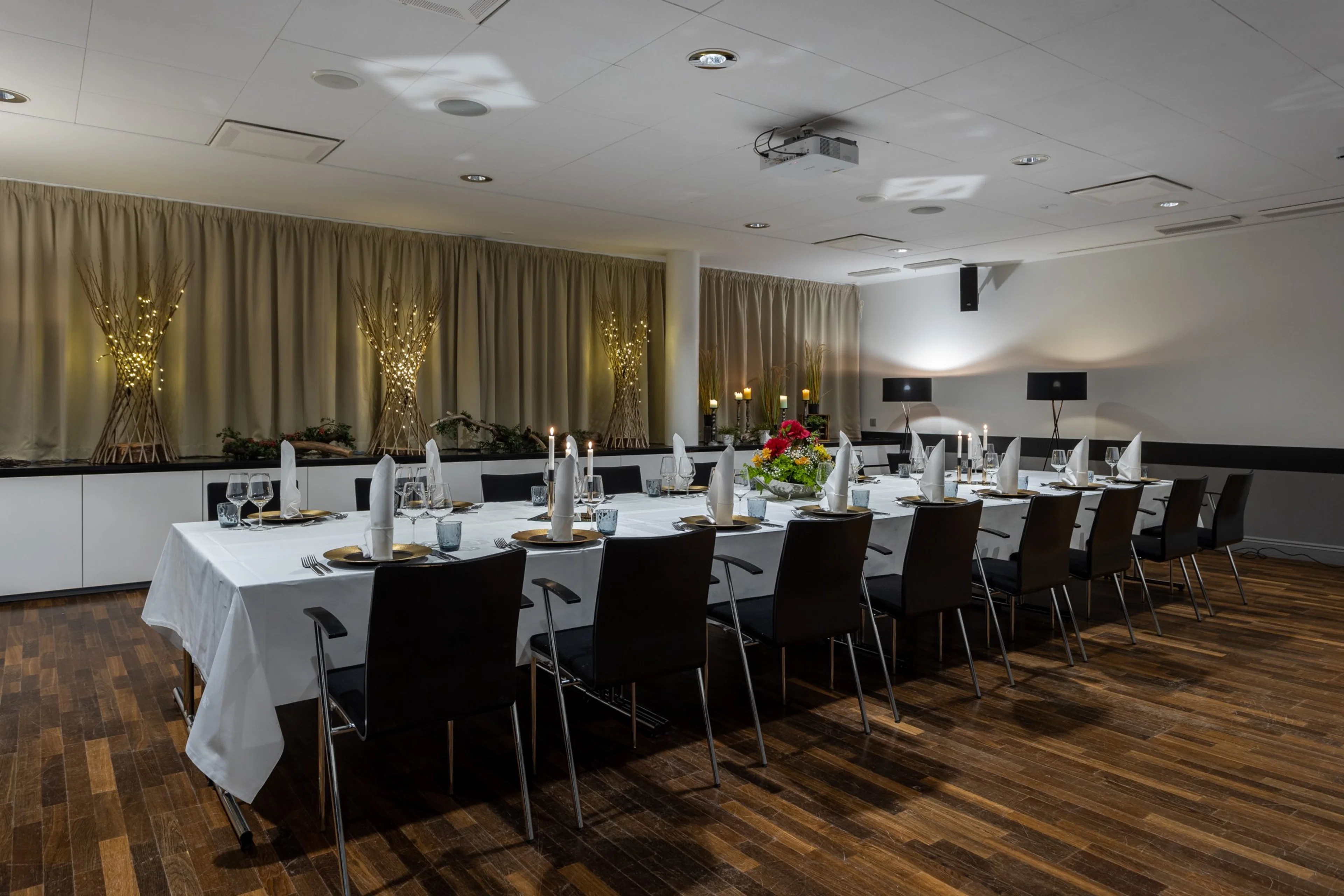 Kokoustila jossa pitkä pöytä valmiiksi katettuna, tummia tuoleja, puinen lattia ja seinillä valaistuja koristeita.