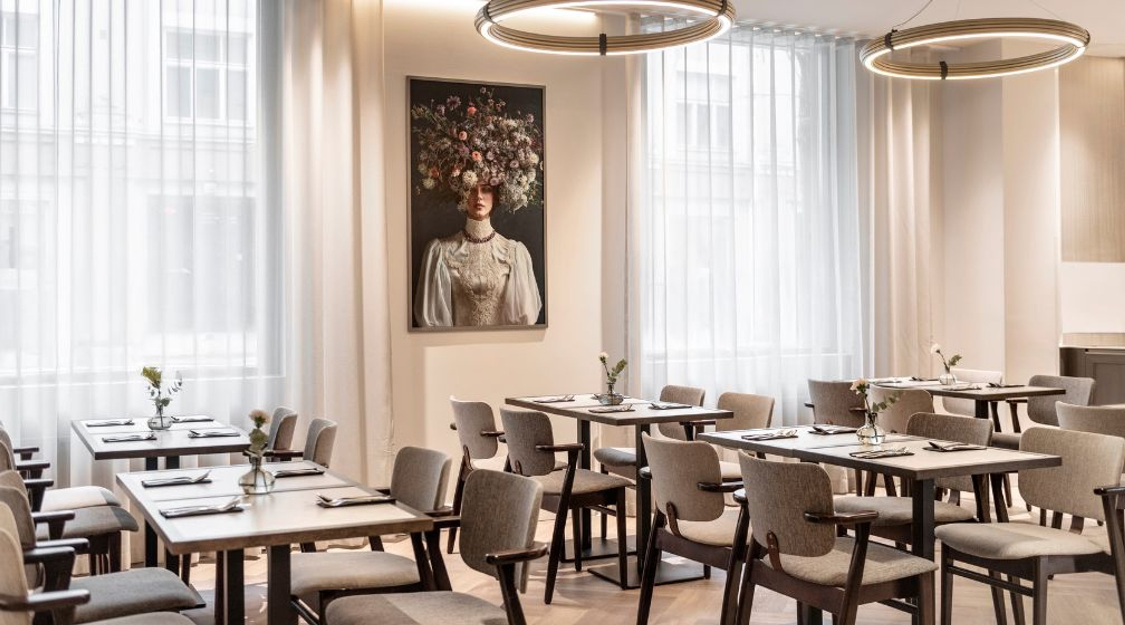 Valoisa ravintola, jossa on vaaleita pöytiä ja tuoleja, koristeellinen taide seinällä ja kaksi pyöreää valaisinta katossa.