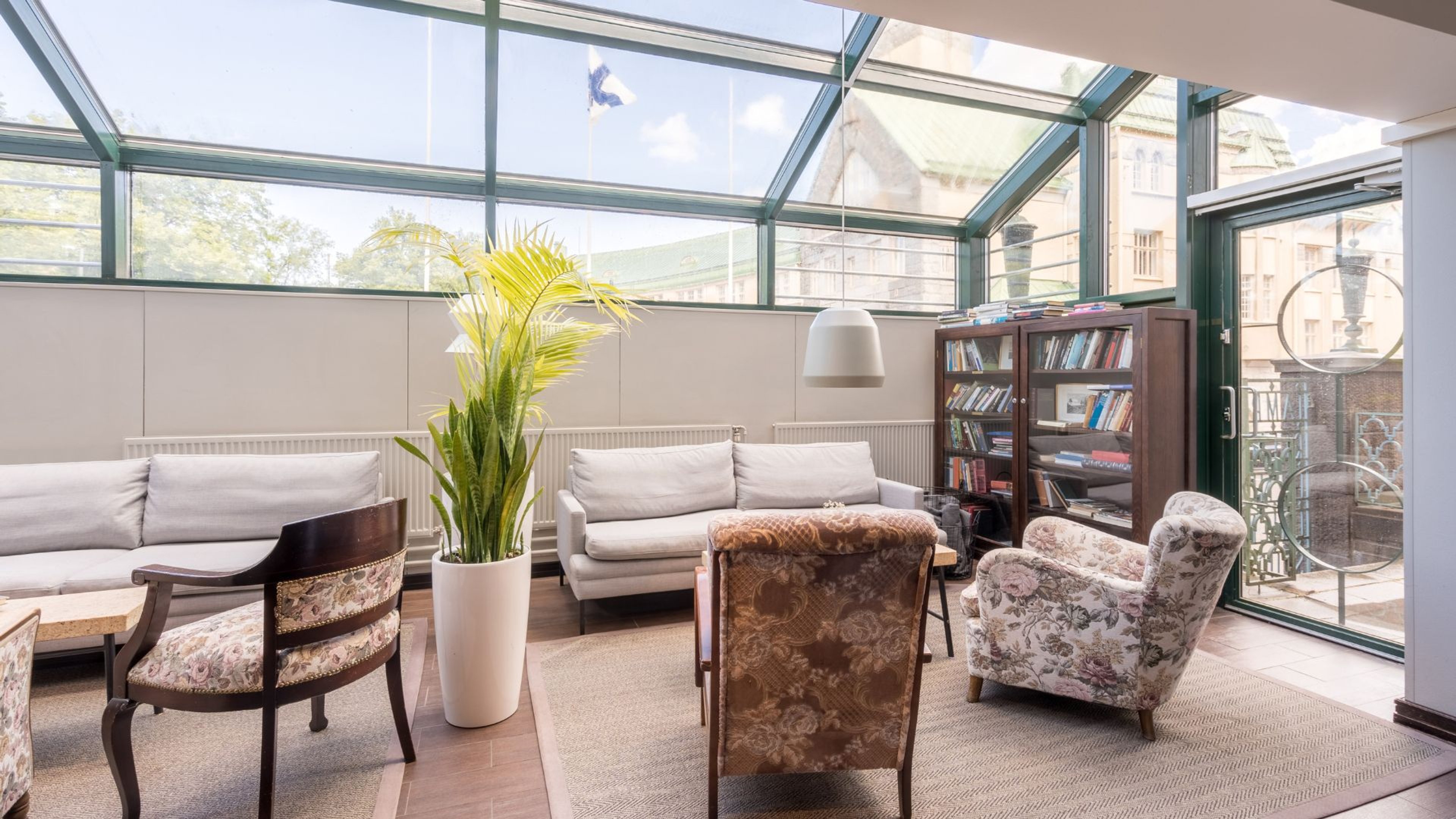 Valoisa lasitettu huone, jossa valkoinen sohva, kuviolliset nojatuolit, kirjahylly ja viherkasvi. Tasainen lattiapinta ja avara tila.