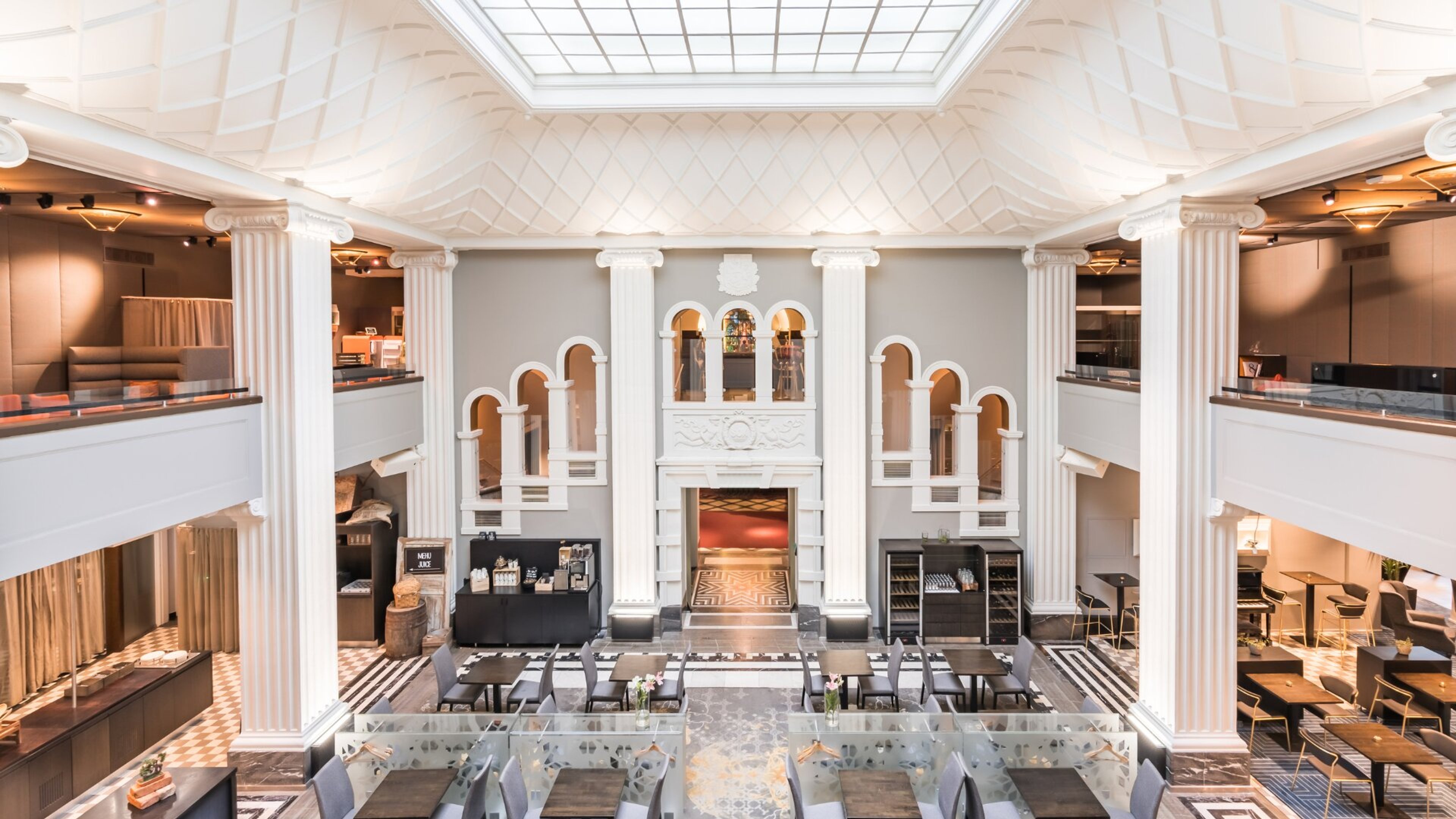 Elegantti sisäpiha klassisilla valkoisilla pylväillä, kattoikkunalla ja kaksikerroksisella ravintola-alueella. Marmorilattialla on useita pöytiä ja tuoleja.