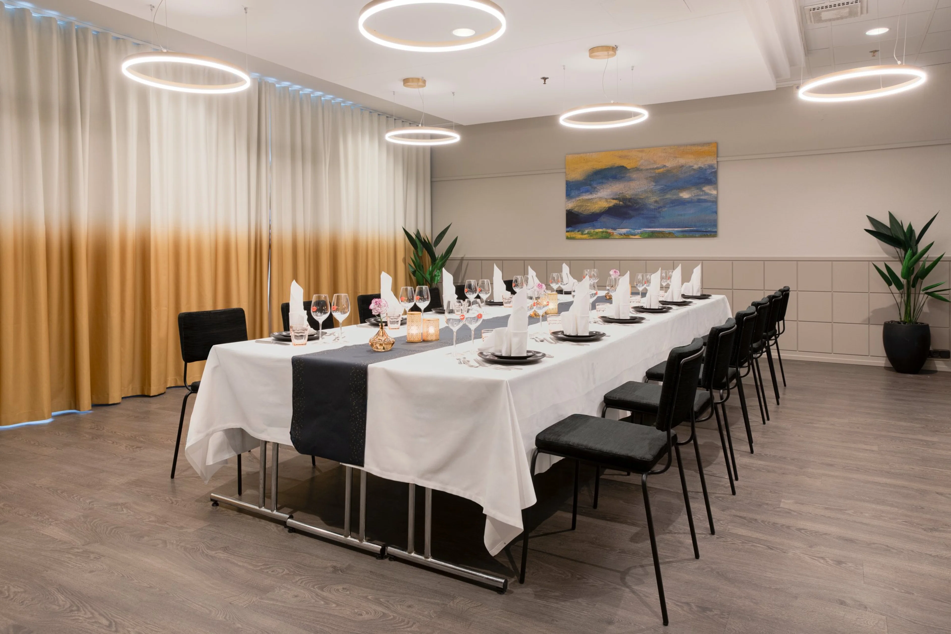 Elegantisti katettu pitkä pöytä juhlatilassa, jossa ympyränmuotoiset valaisimet, ombré-verhot ja taideteos seinällä. Tasainen puulattia.
