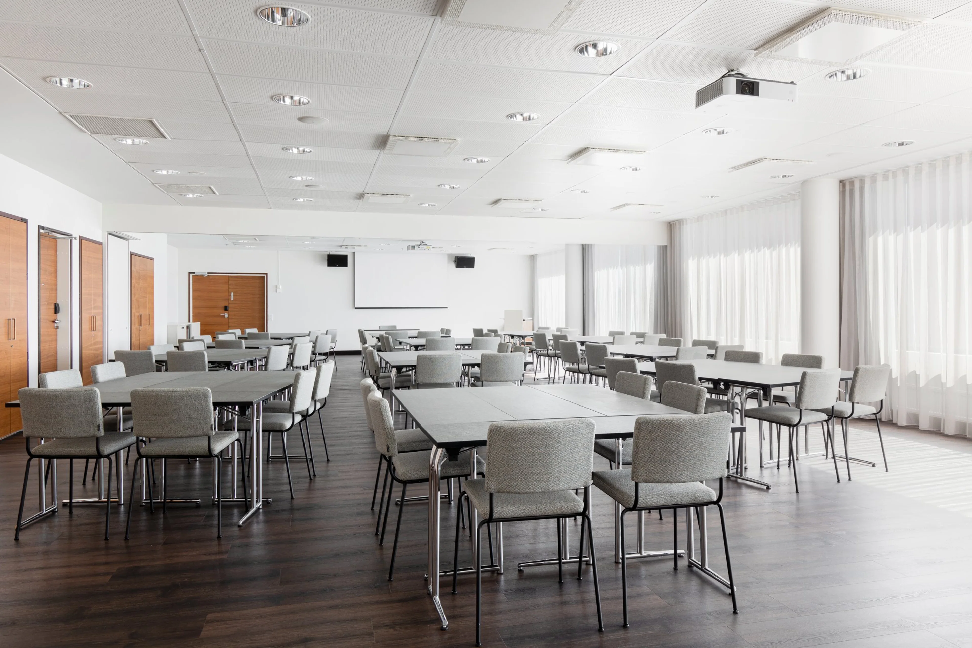 Valoisa kokoushuone pöydillä ja harmailla tuoleilla, puulattia, valkokangas ja projektori, puiset ovet ja ikkunat verhoilla.