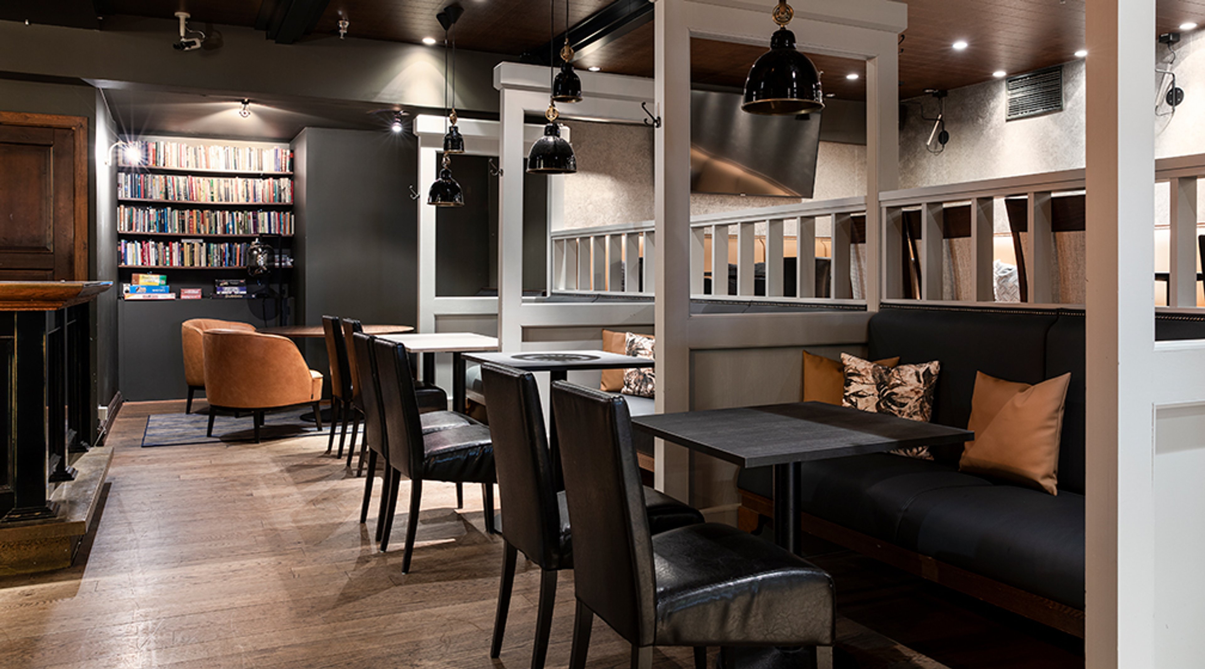 Ravintolan sisätila puulattioilla, jossa mustia pöytiä ja tuoleja, sohvanurkkaus ja kirjahylly. Tilassa roikkuvat mustat valaisimet ja TV-näyttö.