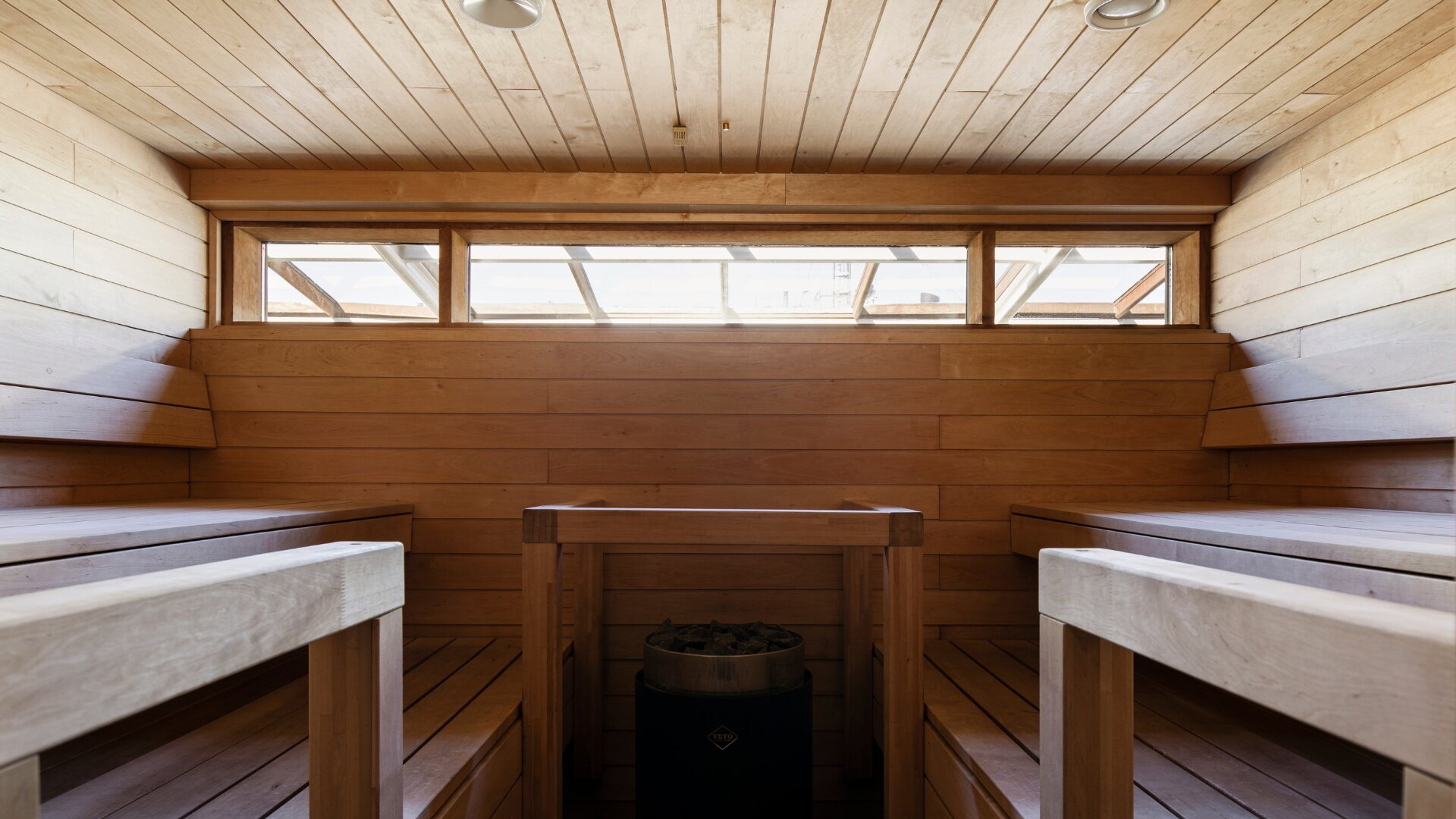 Puupaneloitu sauna, jossa on lauteita molemmin puolin, kiuas keskellä ja pitkä ikkuna tuomassa luonnonvaloa.