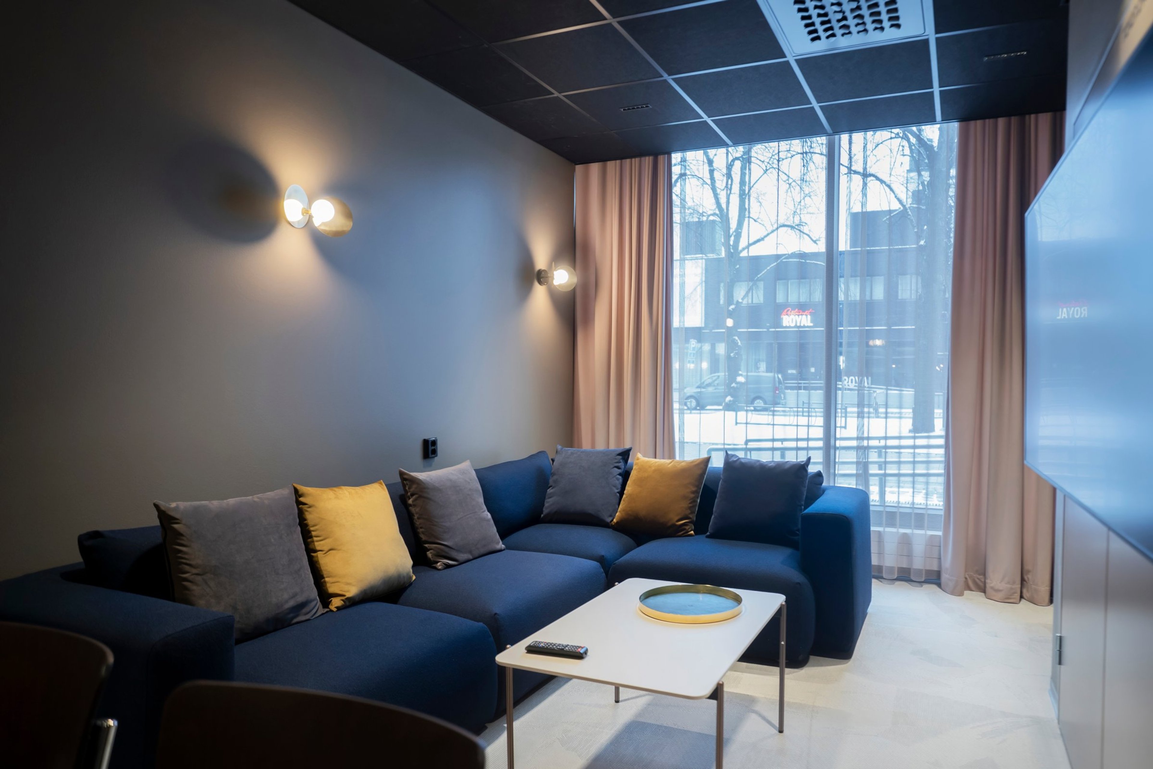 Moderni oleskelutila, jossa sininen sohva värikkäillä tyynyillä, valkoinen sohvapöytä ja suuret ikkunat. Tasainen vaalea lattia, hyvä valaistus.