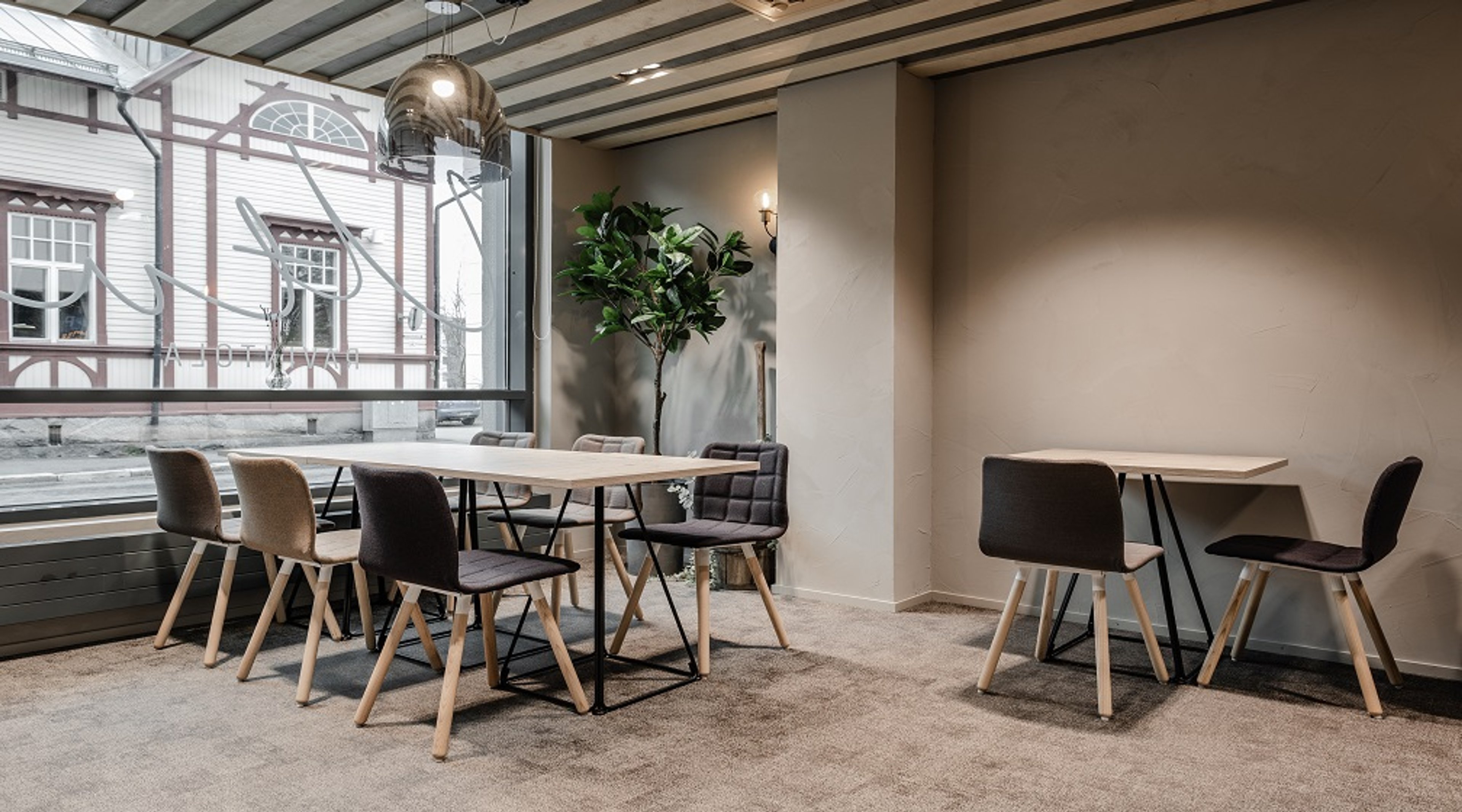 Moderni kokoustila, jossa kaksi pöytää, tuoleja, koristekasveja ja isot ikkunat, joista näkyy vanha puurakennus. Lattia on peitetty matolla.