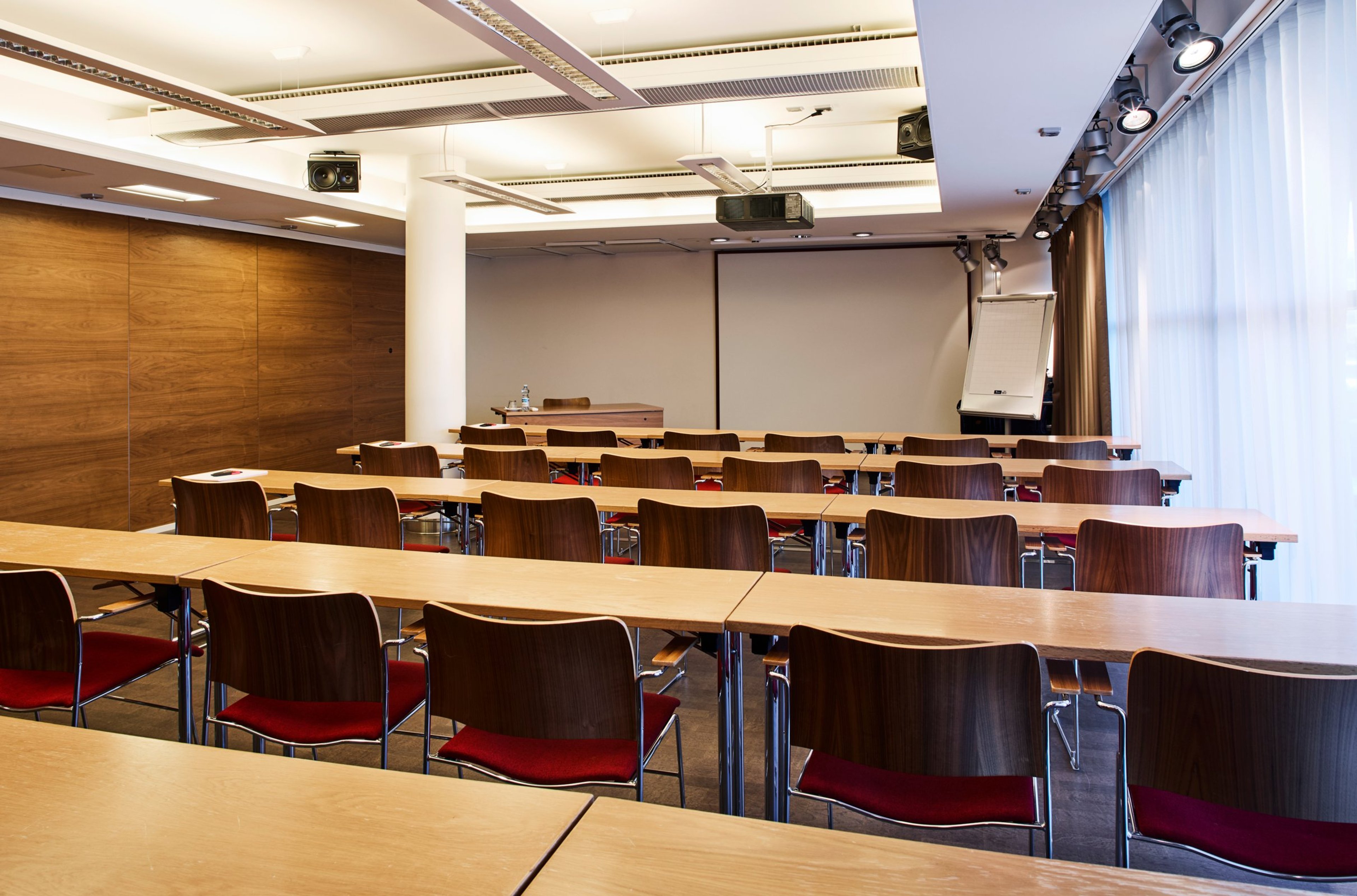 Kokoustila puisilla pöydillä ja tummanpunaisilla tuoleilla, edessä projektori ja valkokangas, seinät puupaneloitu.