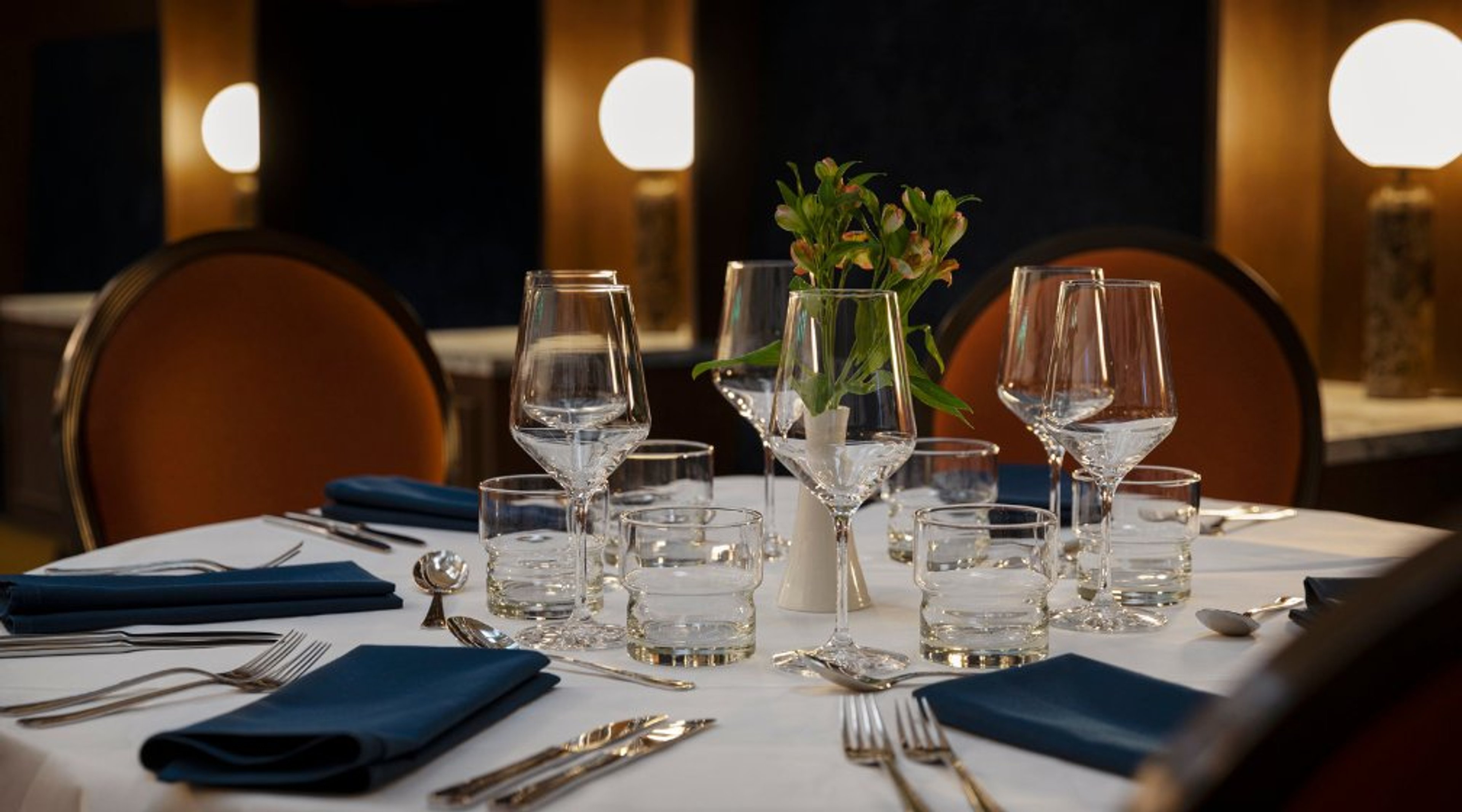 Tyylikkäästi katettu pöytä ravintolassa. Valkoinen pöytäliina, siniset lautasliinat, viinilasit ja pieni kukka-asetelma keskellä.