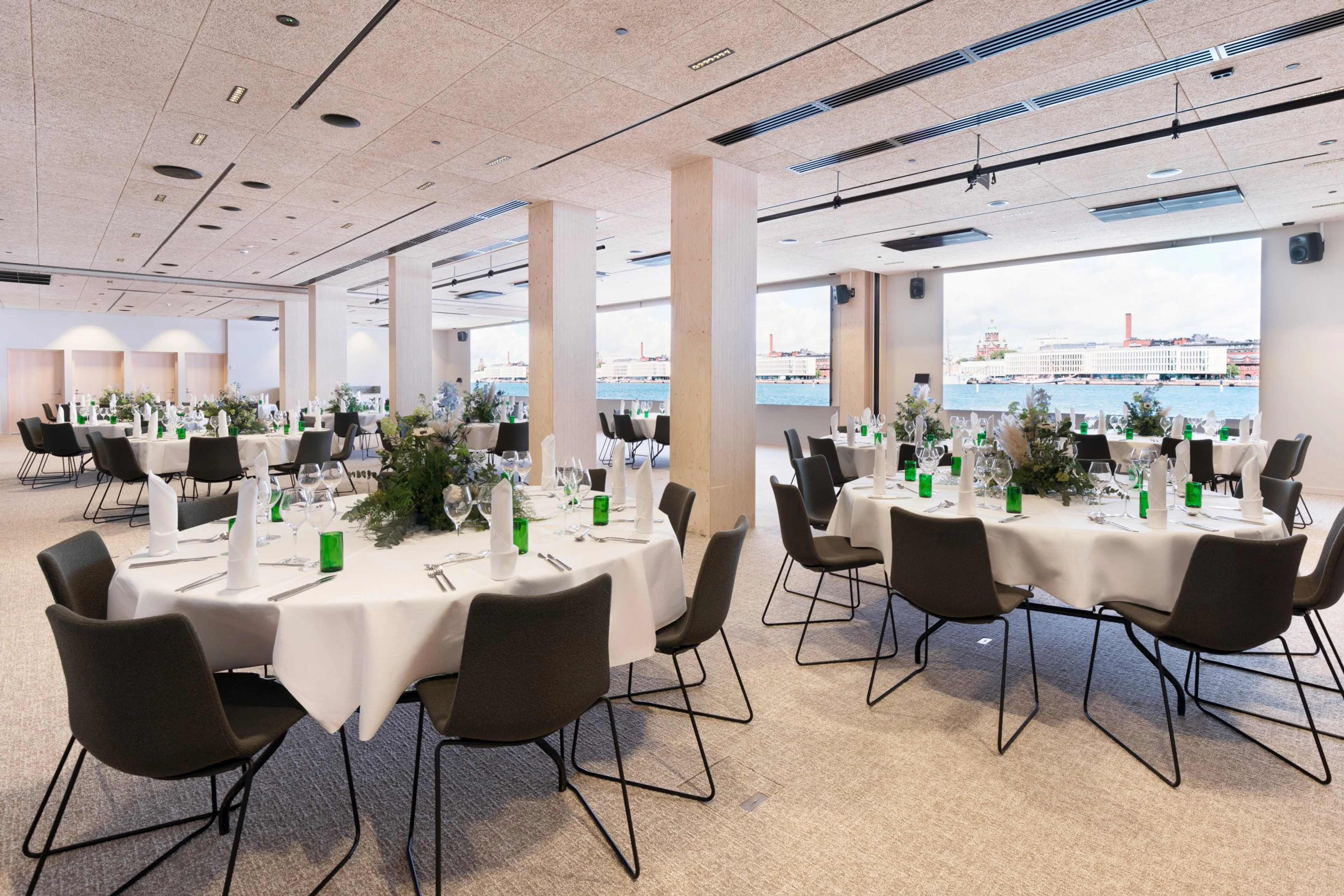 Ljus festlokal med runda bord, vita dukar och svarta stolar. Stora fönster visar havsutsikt. Jämnt golv och rymligt utrymme.