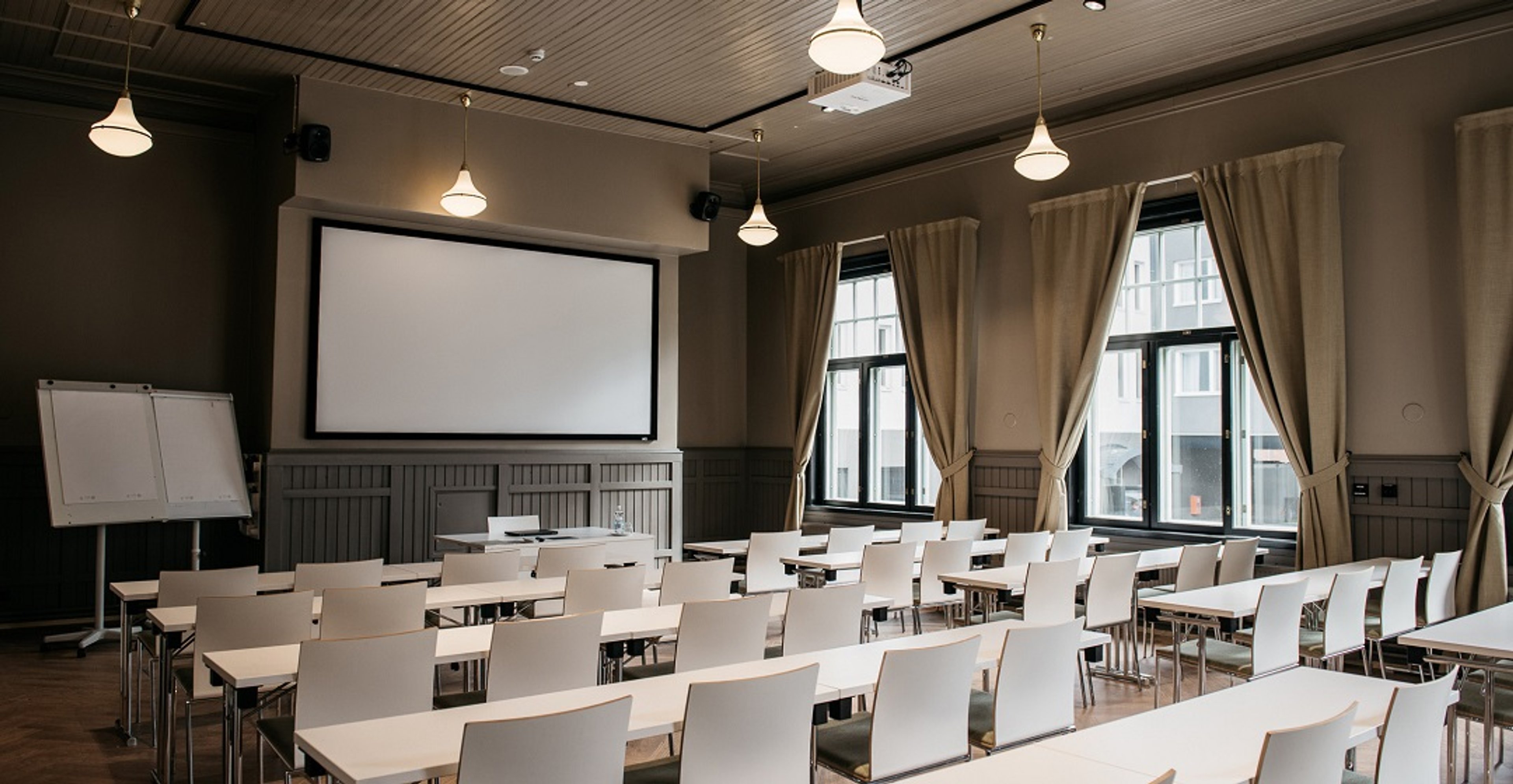 Tilava kokoushuone valkoisten pöytien ja tuolien kanssa. Edessä valkokangas ja fläppitaulu, seinustalla suuret ikkunat beigen verhoin ja katosta riippuvat valaisimet.