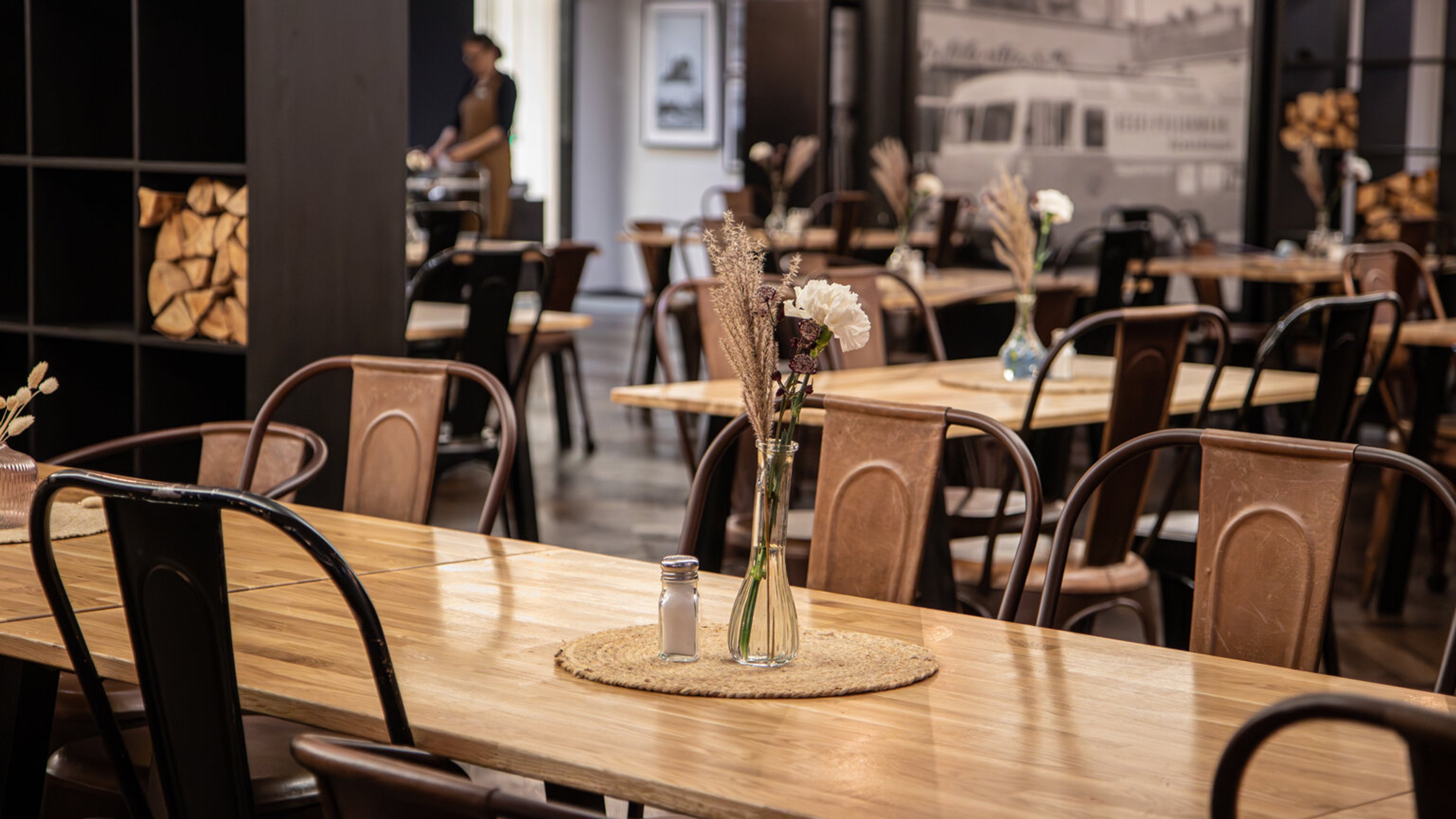 Kahvilan sisustus puisilla pöydillä ja metallisilla tuoleilla. Pöydällä kukkamaljakko ja suolasirotin pyöreällä alustalla.