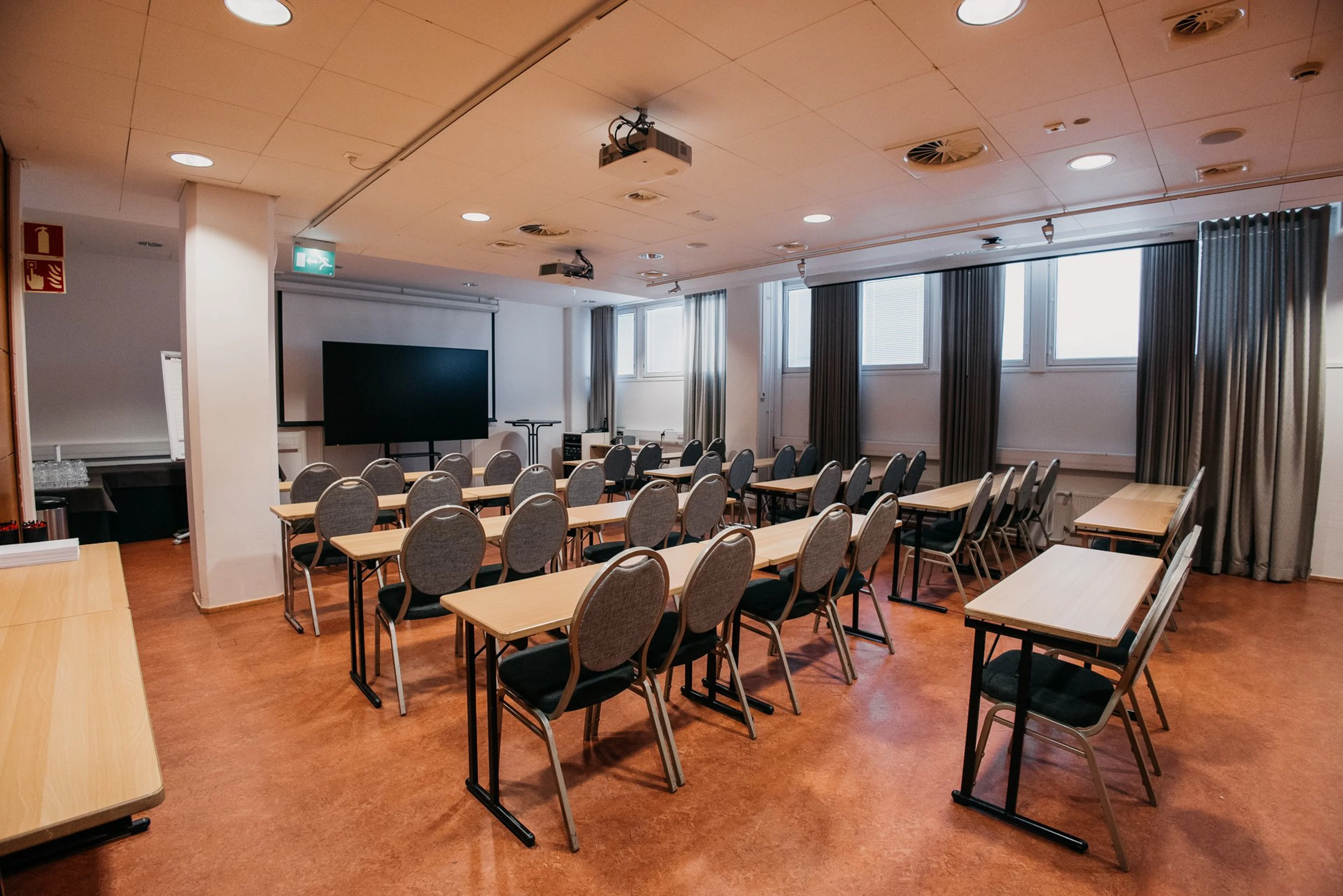 Kokoushuone pöytäriveillä, tuoleilla ja esitysvalkokankaalla. Tilassa projektori katossa, ikkunoita verhoineen ja tasainen lattia.