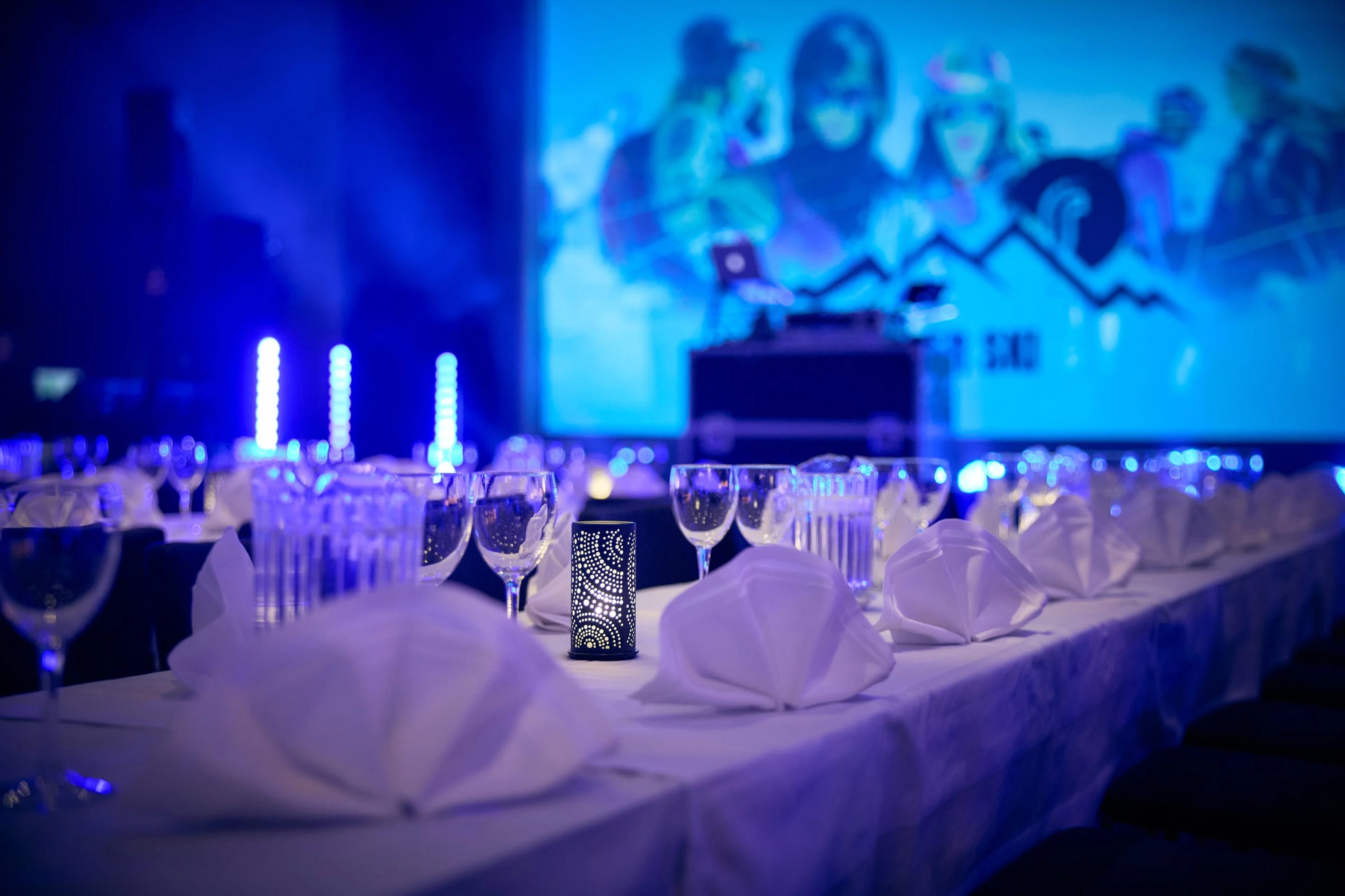 Juhlallisesti katettu pöytä sinisessä valaistuksessa tapahtumaa varten, taustalla näkyy esitysalue ja projektorin sininen valo.