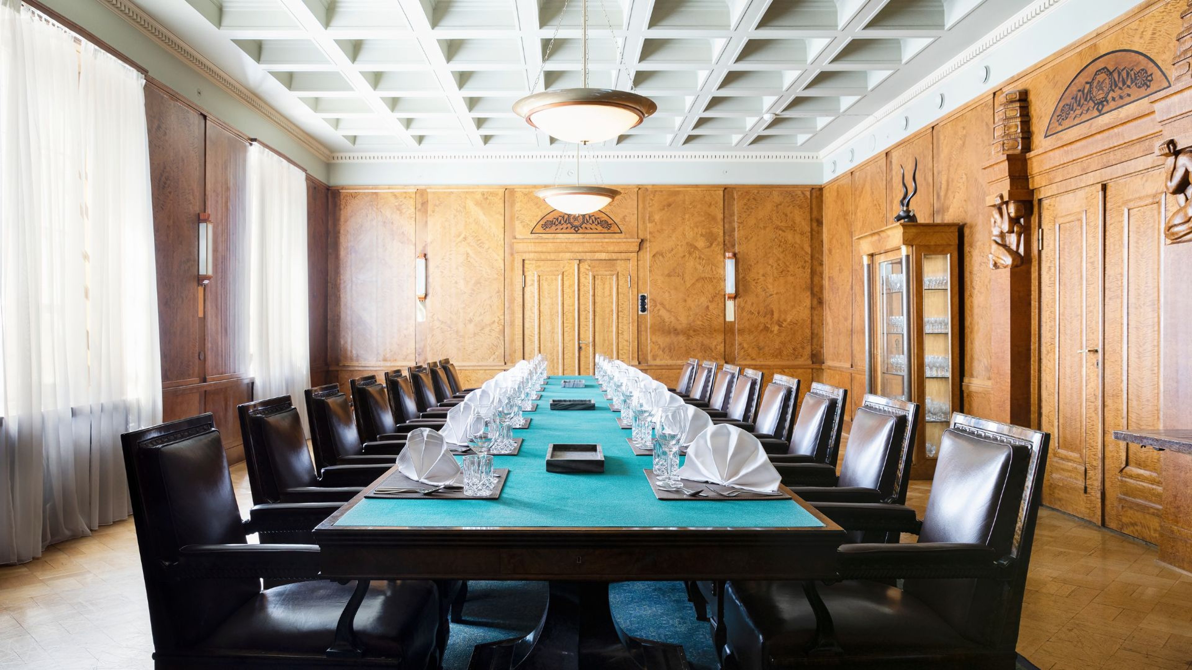 Elegantti kokoushuone puupaneeliseinillä, pitkä pöytä turkoosilla pöytäliinalla, tummat nahkatuolit ja koristeellinen kattovalaisin.