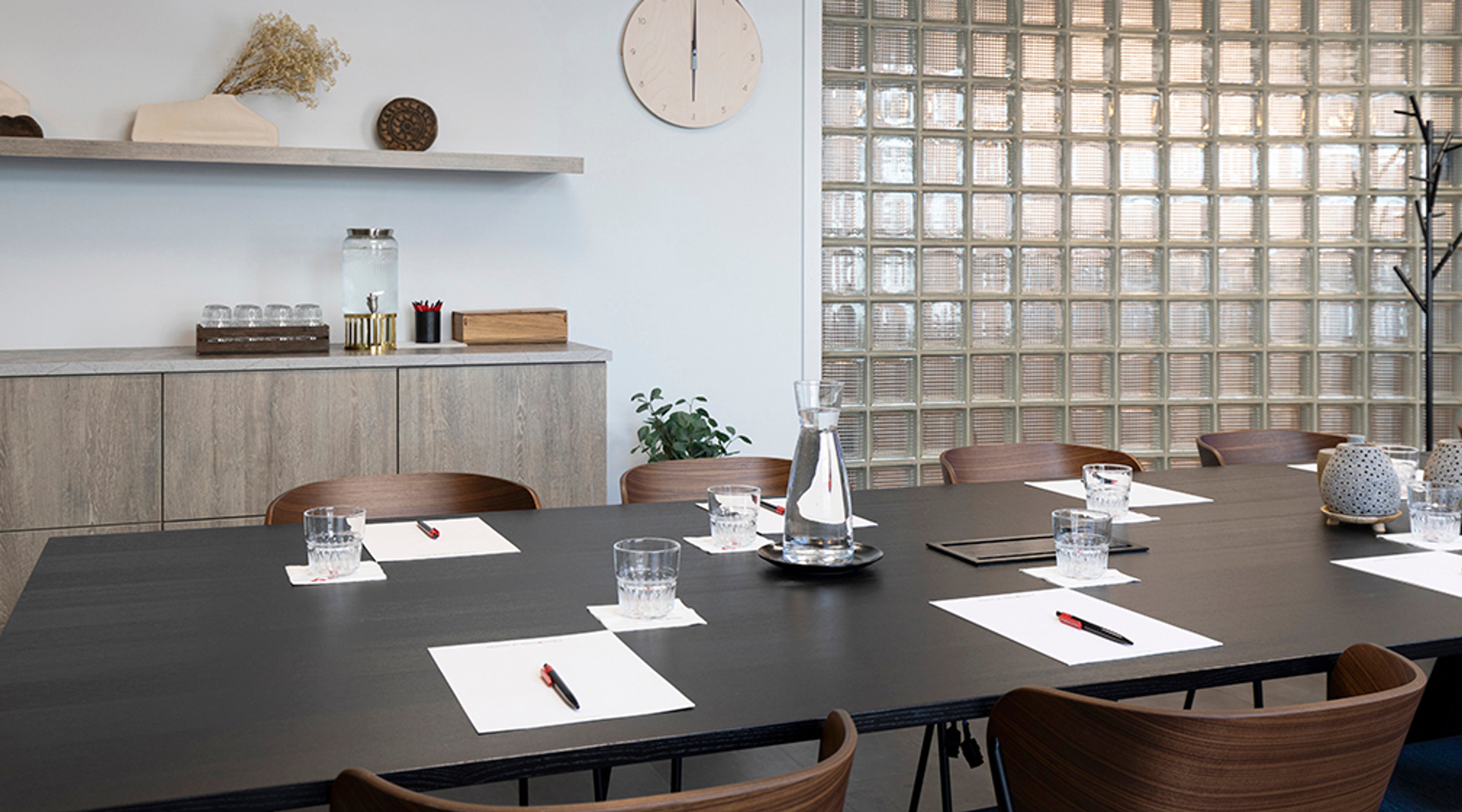 Moderni neuvotteluhuone, jossa puinen pöytä, muistilehtiöt, vesikannu ja lasit. Taustalla lasitiiliväliseinä ja pieni keittiönurkkaus.