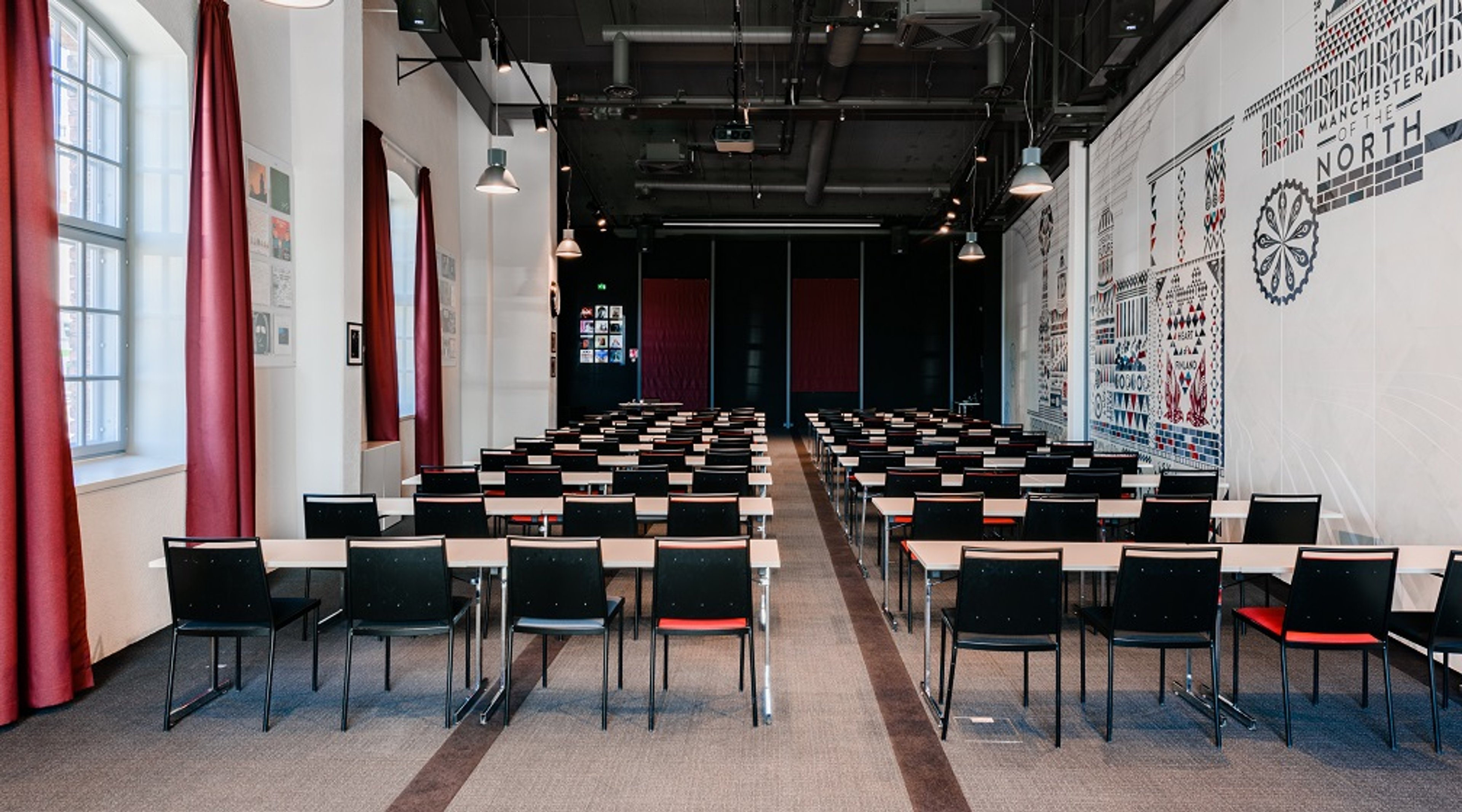 Moderni kokoustila, jossa on riveittäin mustia tuoleja ja vaaleita pöytiä, punaisia verhoja ikkunoilla ja graafinen taideteos seinällä.