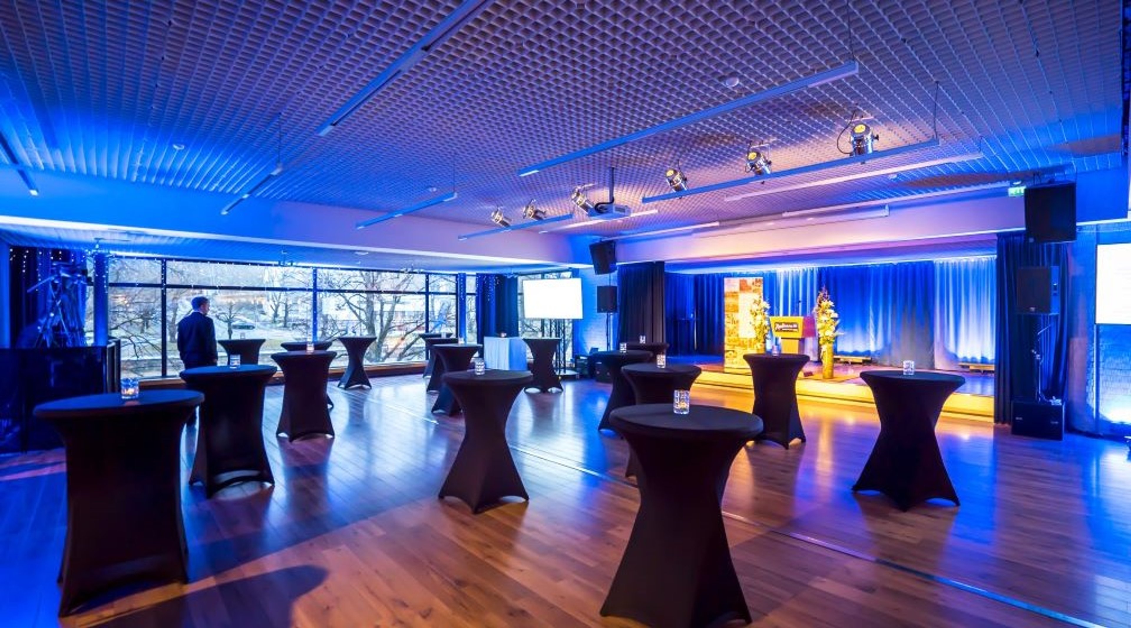 Tilava tapahtumatila sinisessä valaistuksessa. Korkea katto, puulattia ja korkeita cocktailpöytiä mustilla liinoilla. Suuri ikkunaseinä ja lava sinisellä verholla.