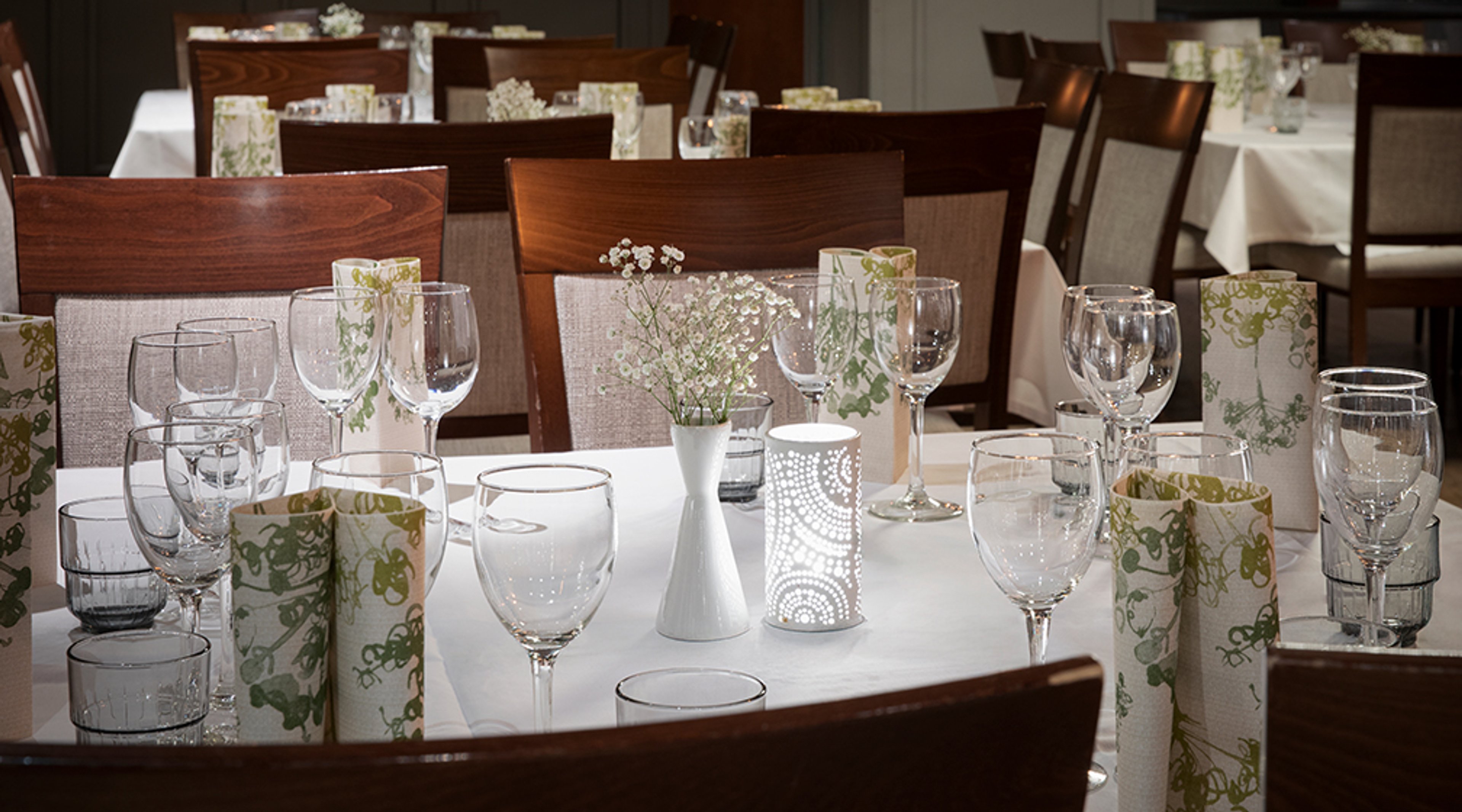 Tyylikkäästi katettu ravintolan pöytä, jossa viinilaseja, koristeellisia kynttilänjalustoja ja pieniä kukka-asetelmia valkoisilla pöytäliinoilla.