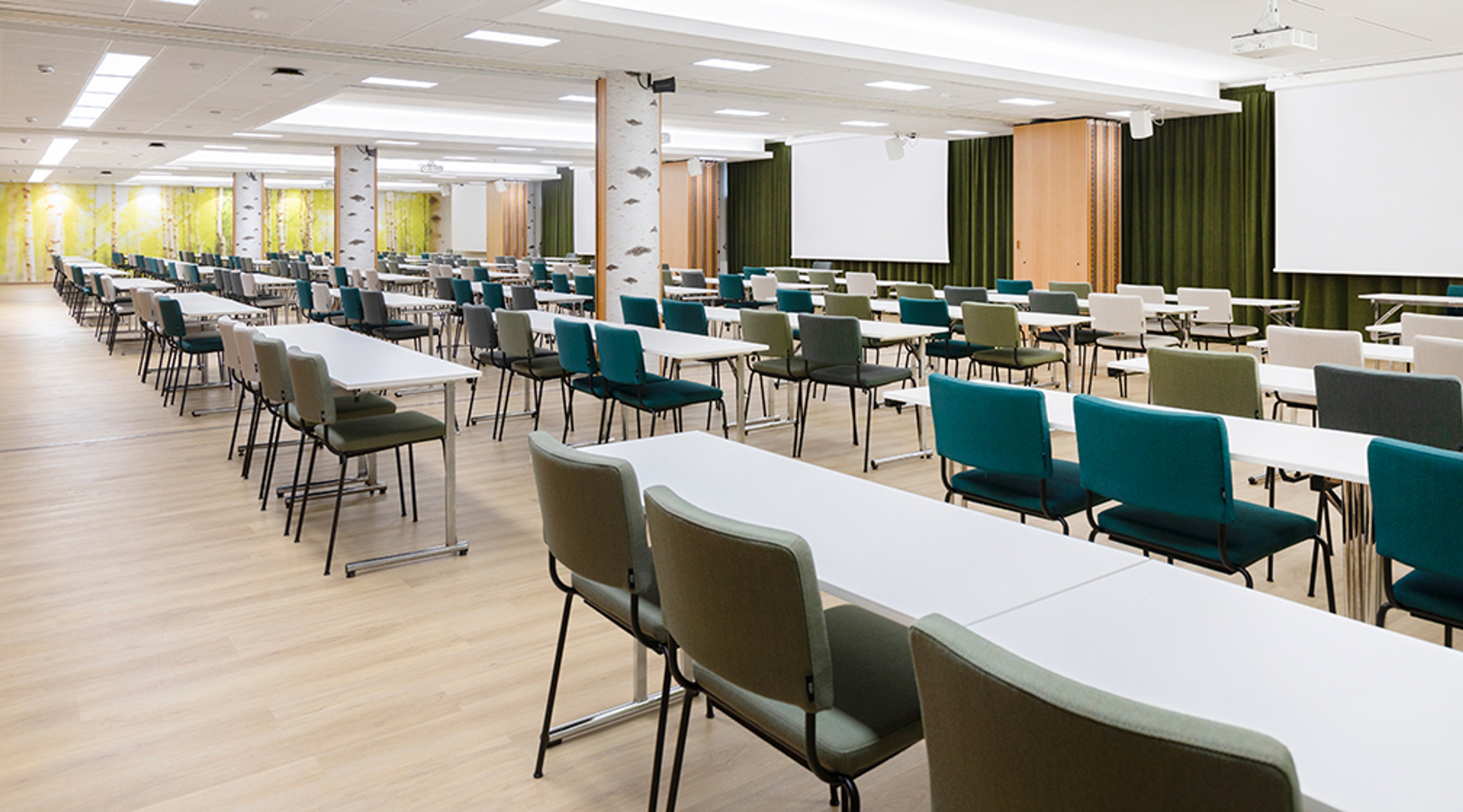 Avara konferenssihuone pitkillä pöydillä ja sinisillä sekä harmailla tuoleilla. Edessä valkokangas ja vihreät verhot. Vaalea lattia ja hyvä valaistus.