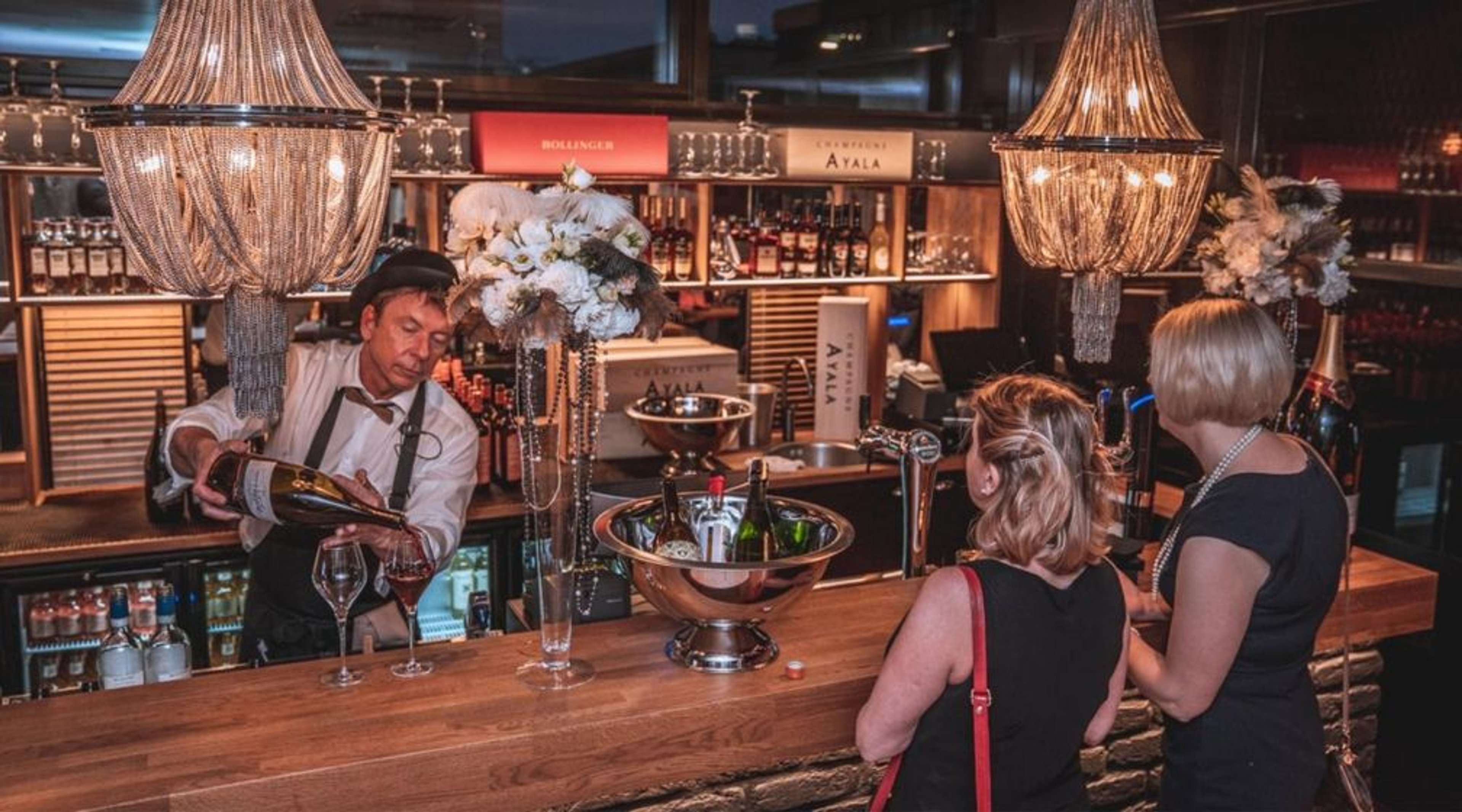 Tunnelmallinen baaritiski, jossa elegantit kattokruunut. Baarimikko kaataa samppanjaa, pöydällä jäähdytysastiat ja kukka-asetelmat.