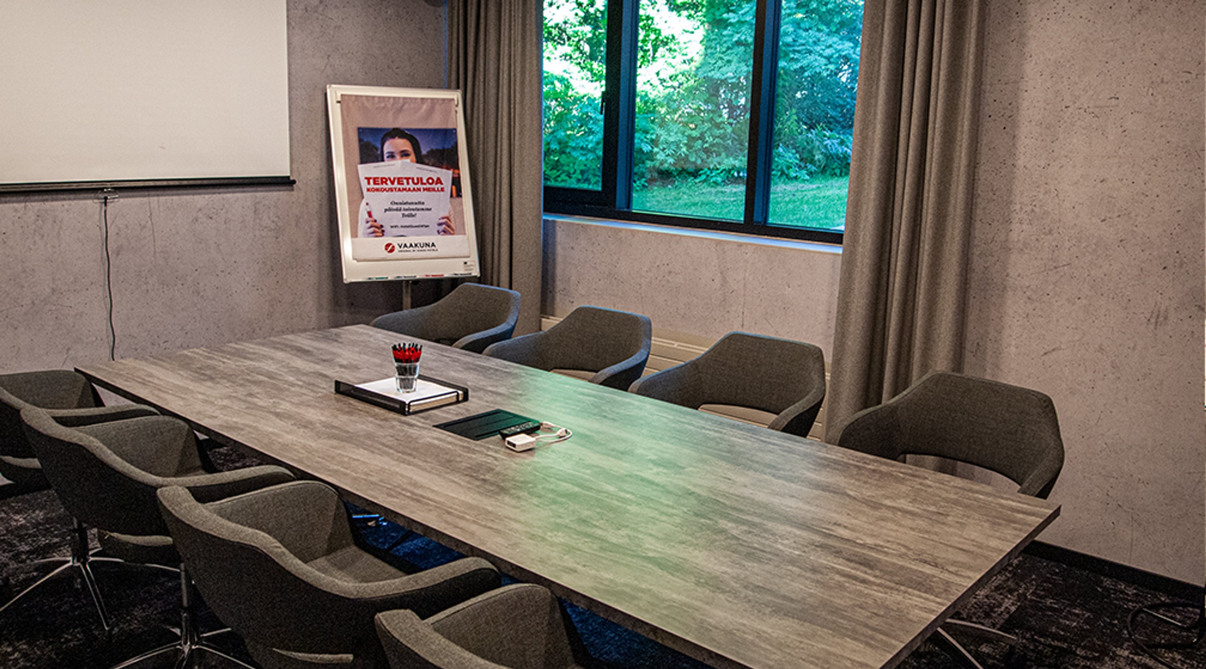 Kokoushuone jossa puinen pöytä ja harmaat tuolit, suuret ikkunat puistomaisemaan, projektorikangas ja tervetuloviesti.