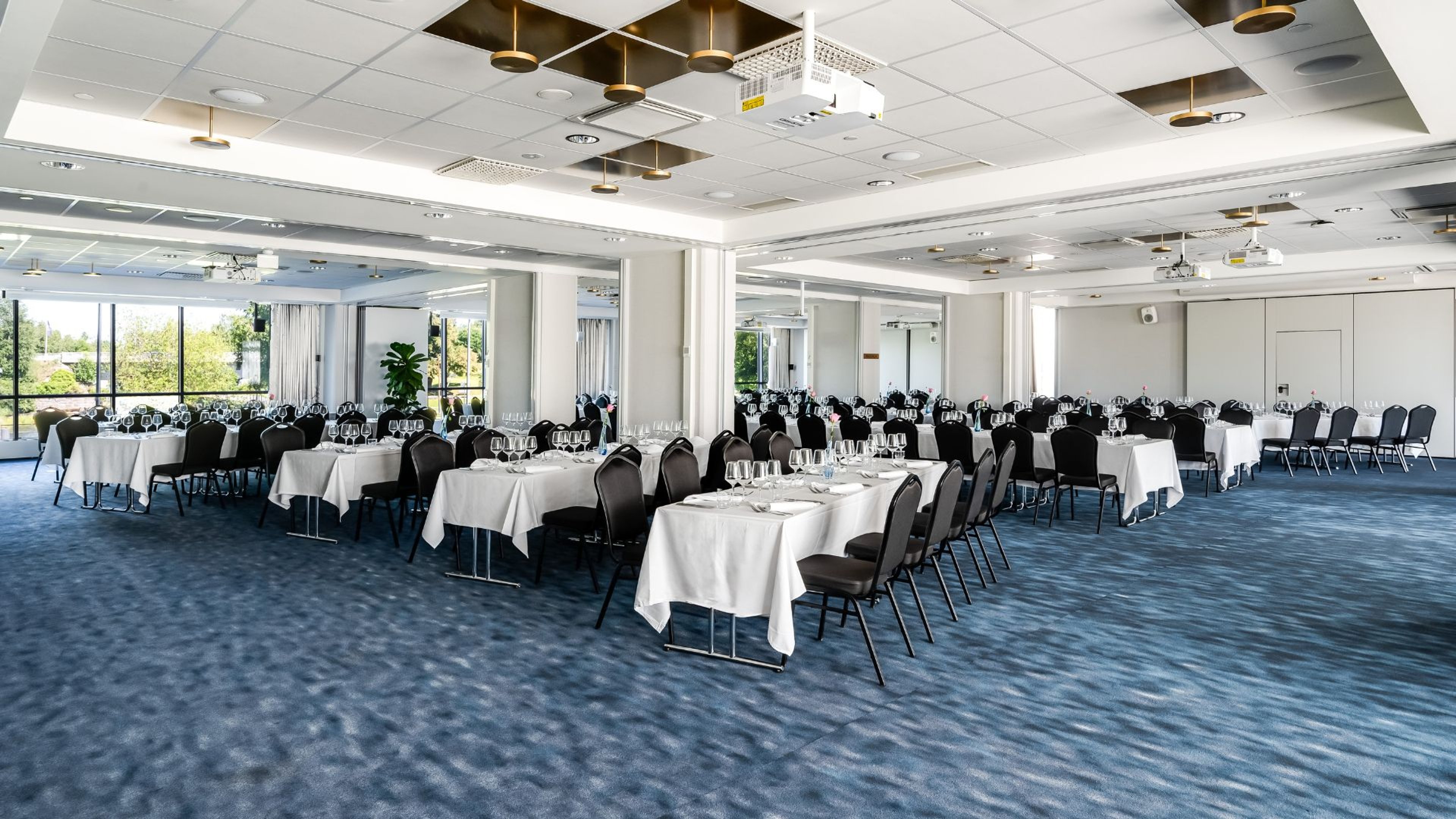 Avara juhlatila, jossa useita pöytiä valkoisilla liinoilla ja mustilla tuoleilla. Sininen kokolattiamatto, suuret ikkunat ja videotykki katossa.