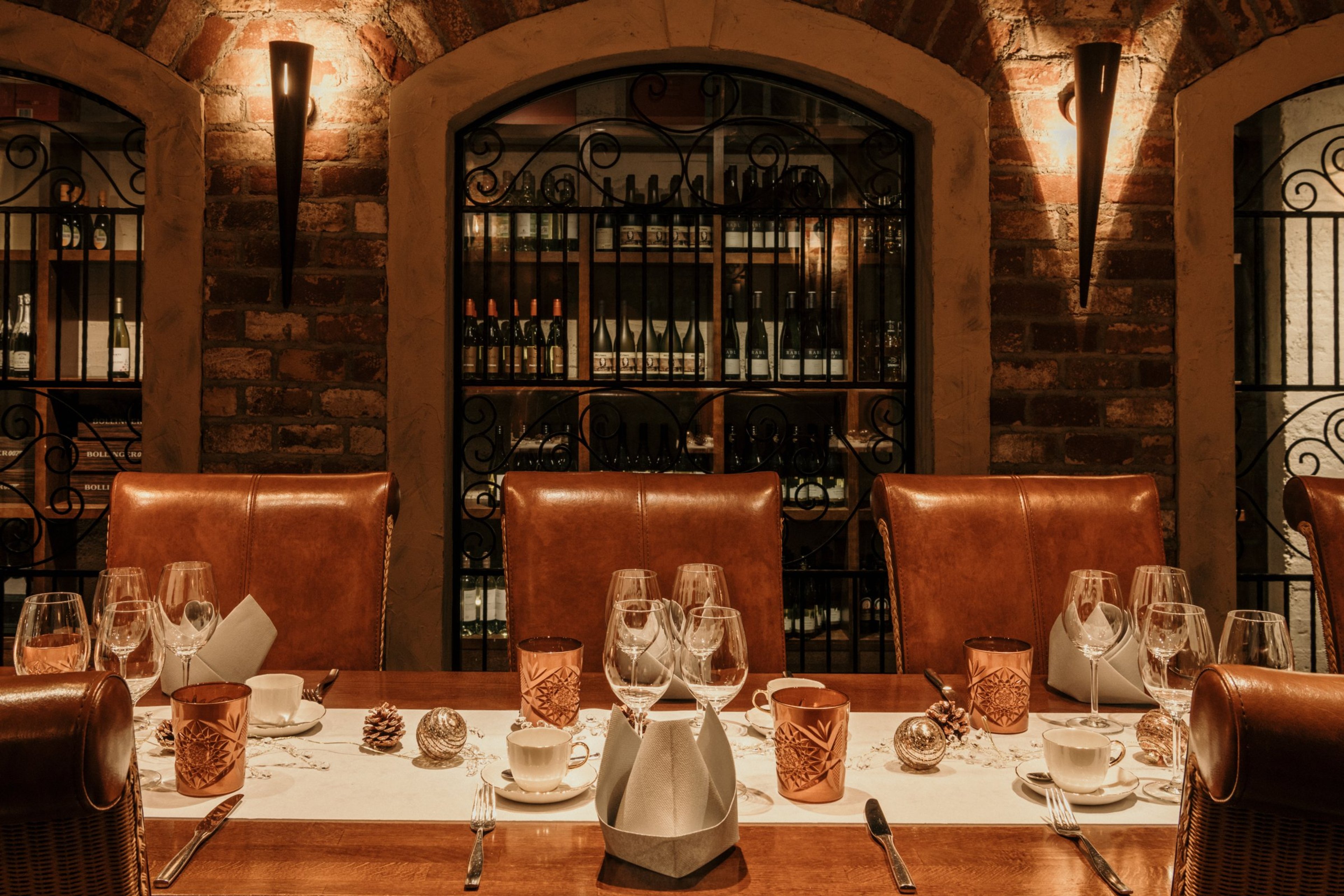 Tunnelmallinen viinikellaritila tiiliseinineen, jossa elegantti ruokapöytä nahkatuoleilla ja kattaus. Taustalla rautaristikkojen takana viinipulloja.
