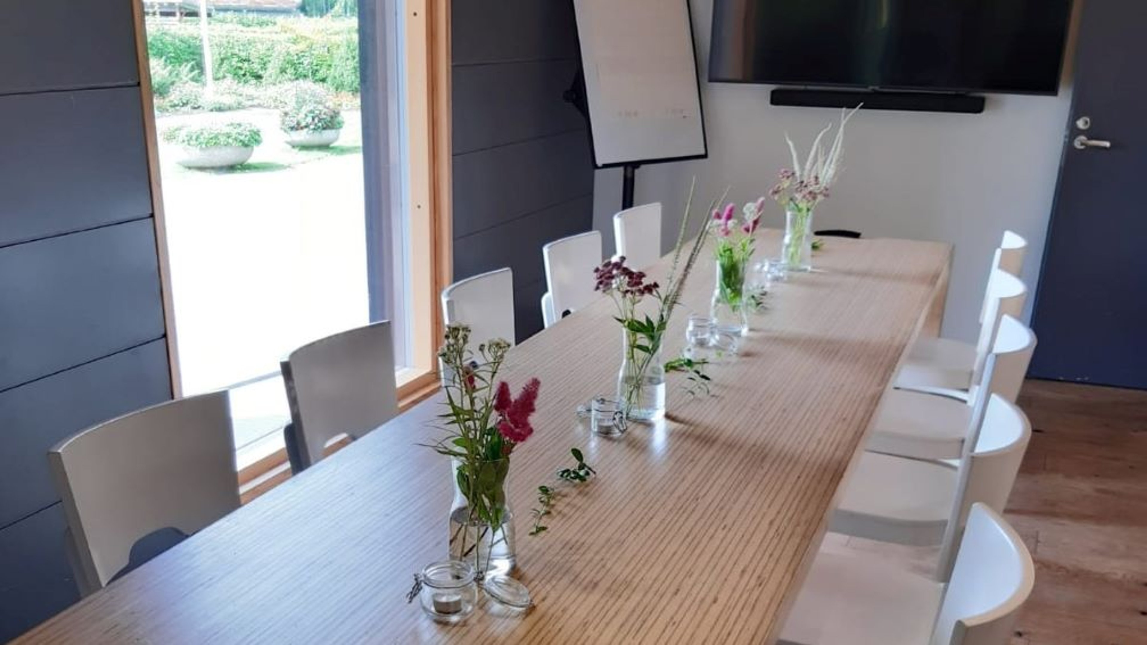 Kokoushuone pitkällä puisella pöydällä, valkoisilla tuoleilla ja pienillä kukka-asetelmilla. Ikkunasta näkyy vihreä puutarha.