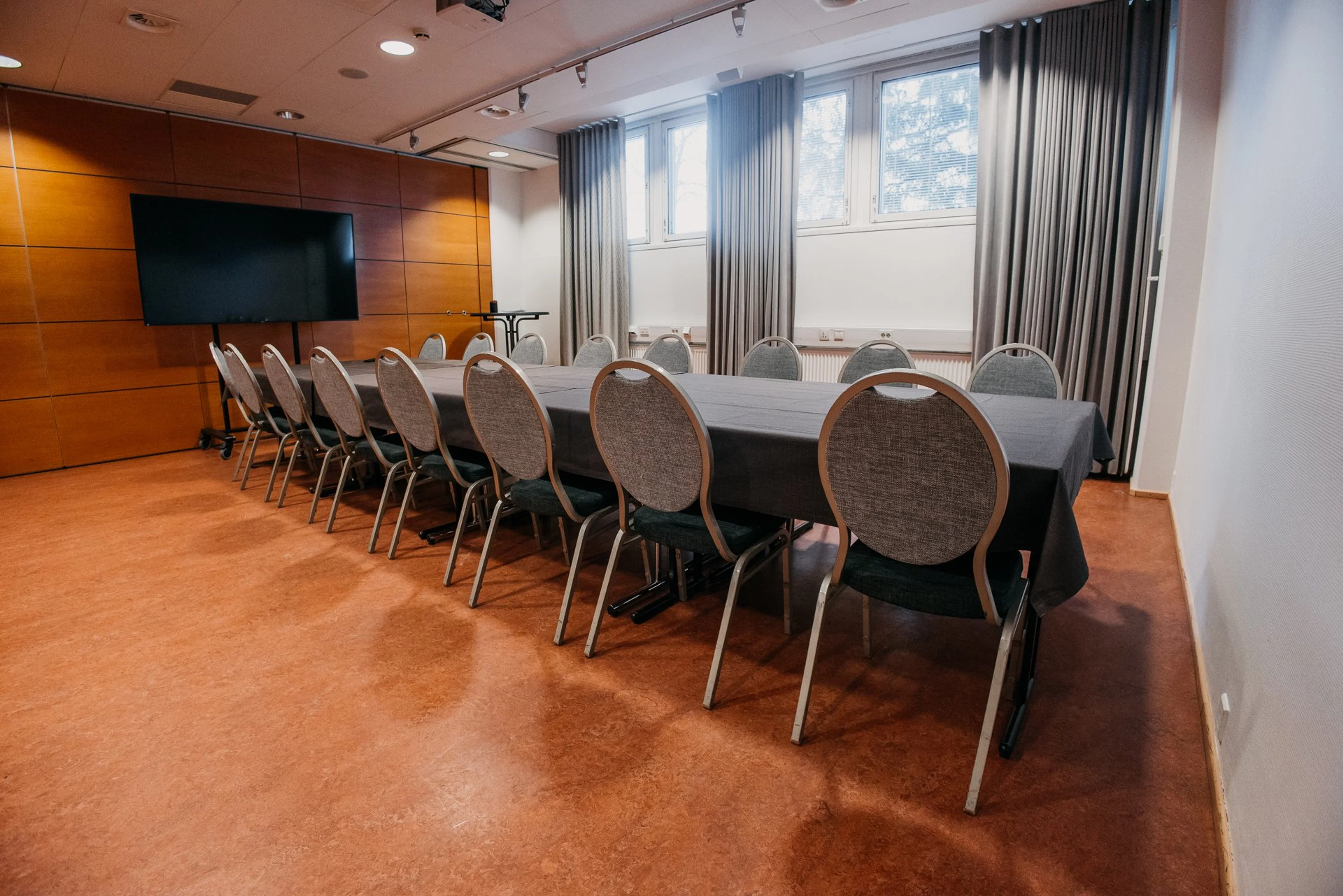 Kokoushuone, jossa on pitkä pöytä mustalla liinalla, harmaat tuolit, televisio puupaneeliseinällä ja tasainen kova lattia.